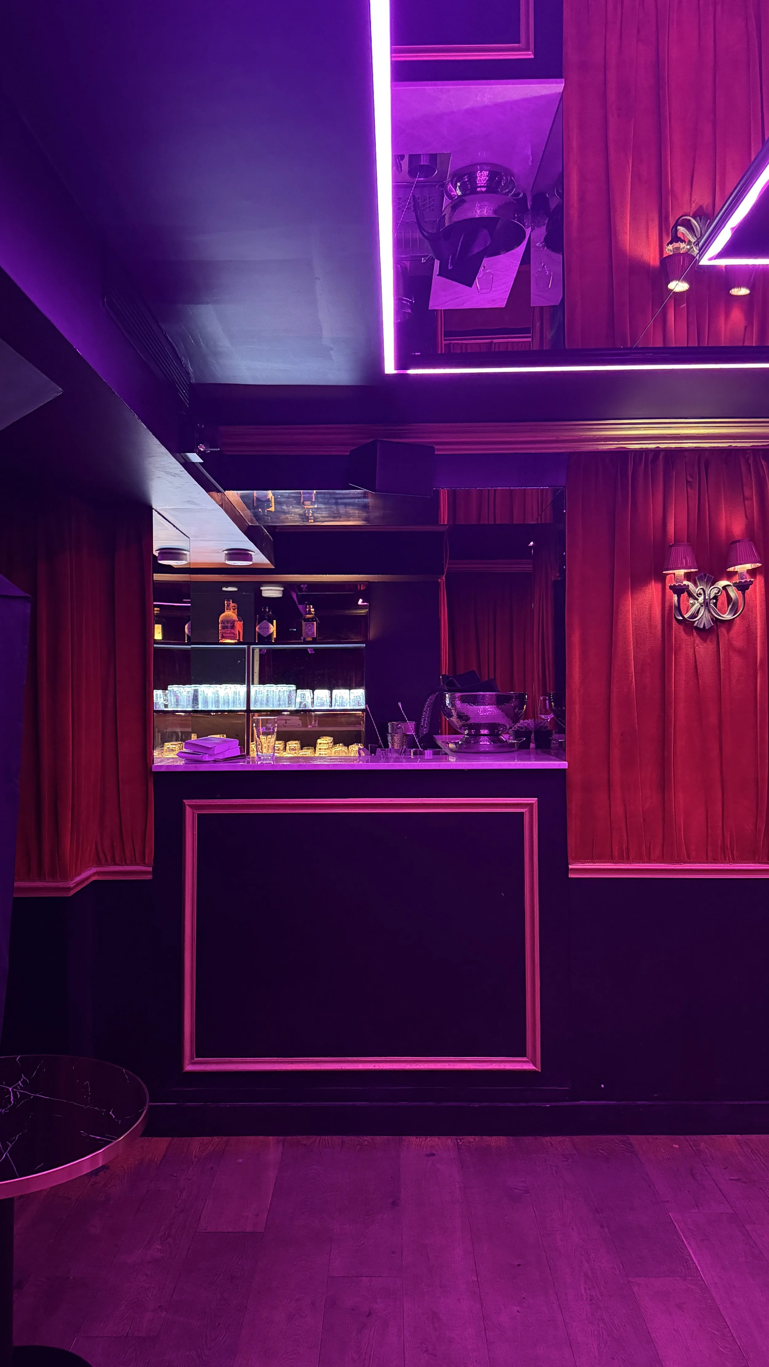 A woman prepares drinks in a bar lit by red and purple lights, with glasses arranged on the counter and bottles of alcohol in the background.