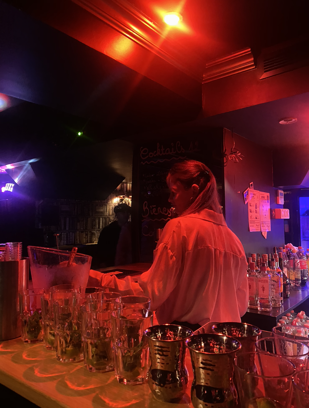 Une femme prépare des boissons dans un bar éclairé par des lumières rouges et violettes, avec des verres disposés sur le comptoir et des bouteilles d'alcool en arrière-plan.