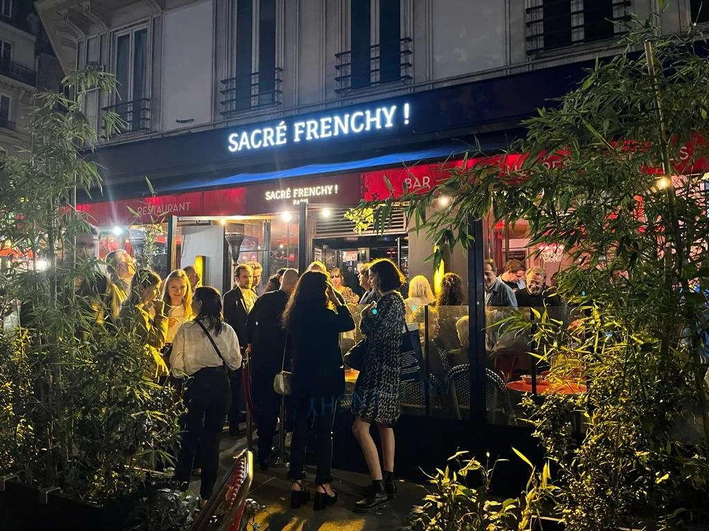 A crowd of people stands outside a restaurant/bar calledSacré Frenchy!" at night, surrounded by green plants and under neon signs.