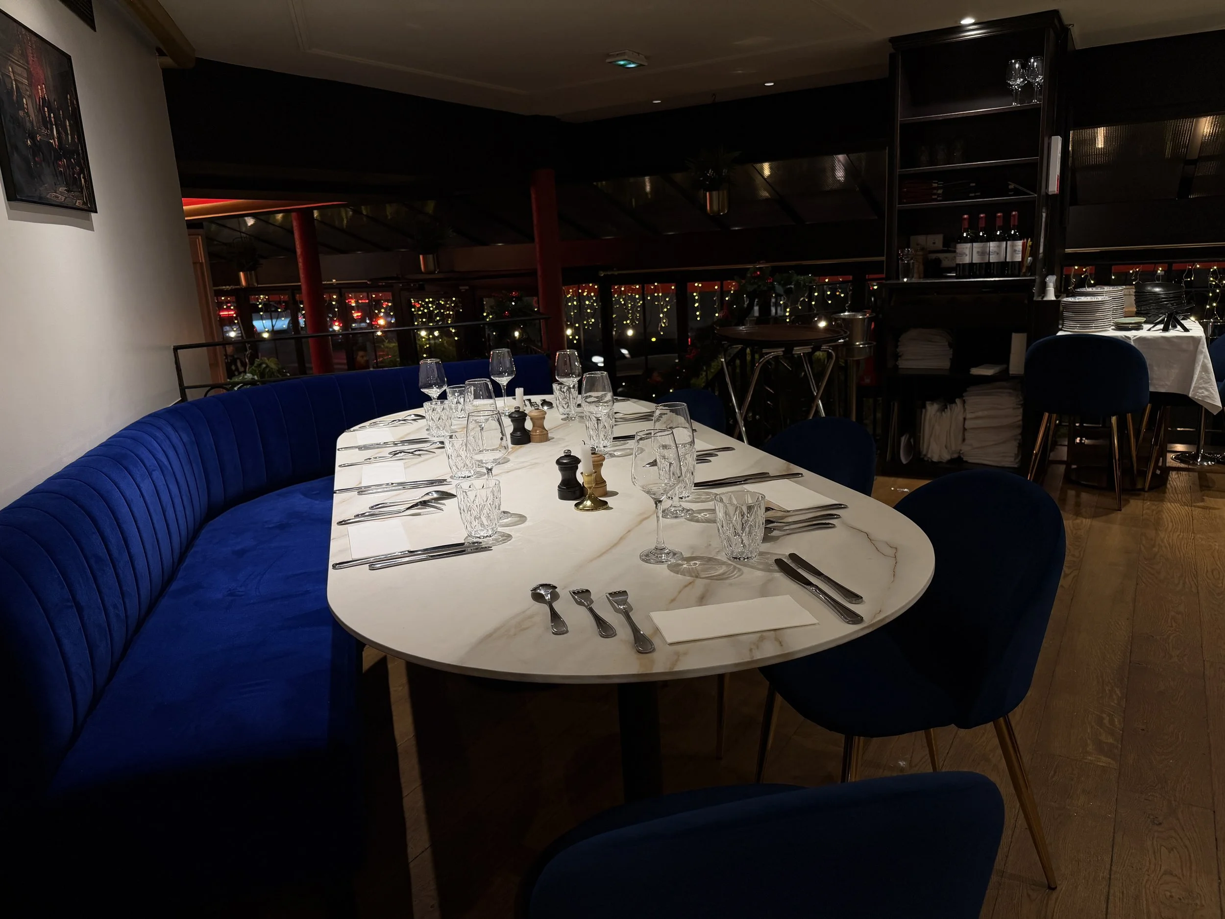 Elegant restaurant interior with well-laid tables, white tablecloths and wooden chairs. Candelabras adorn some tables. The walls are decorated with paintings and the ceiling has an intricate pattern. The lighting is soft and warm.