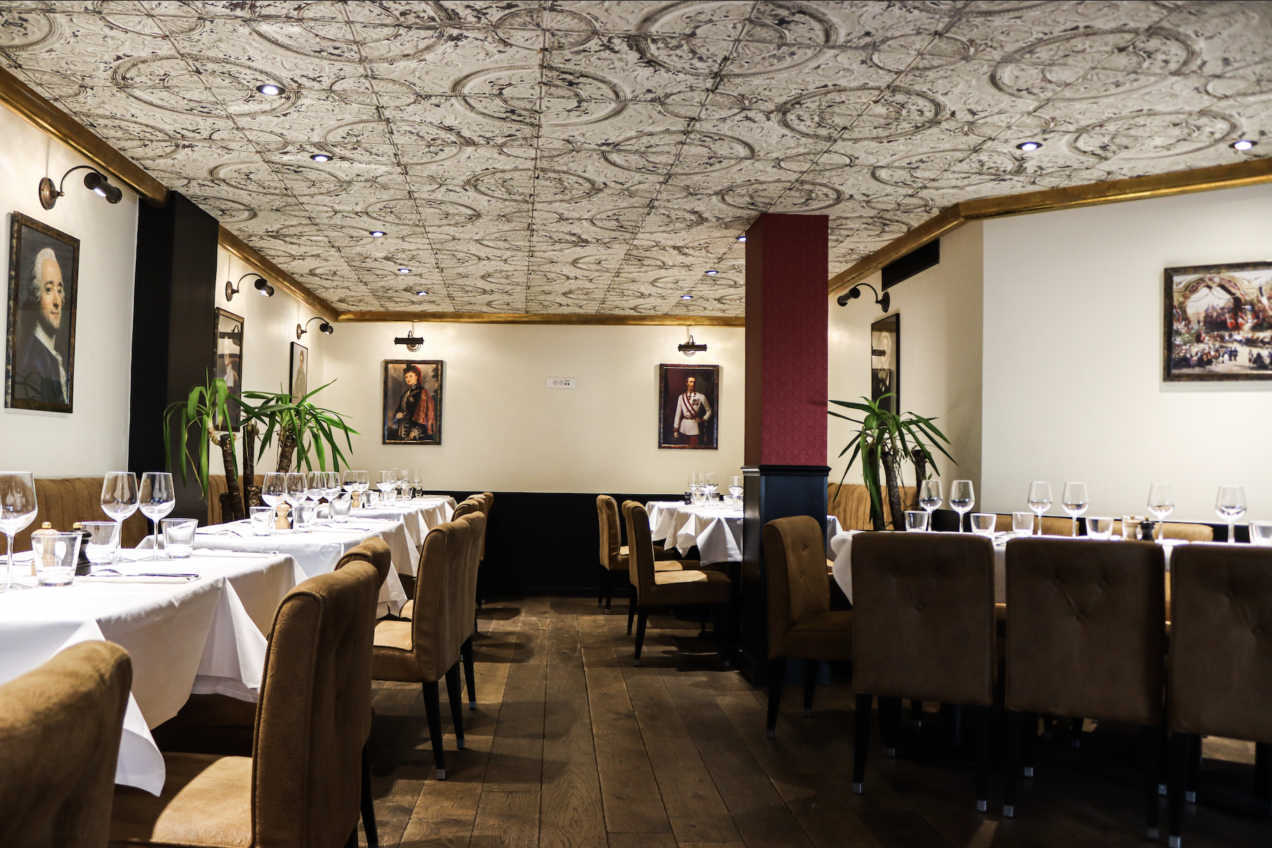 Intérieur d'un restaurant élégant avec des tables dressées, des chaises marron, des nappes blanches, des verres à vin, des plantes et des tableaux sur les murs.