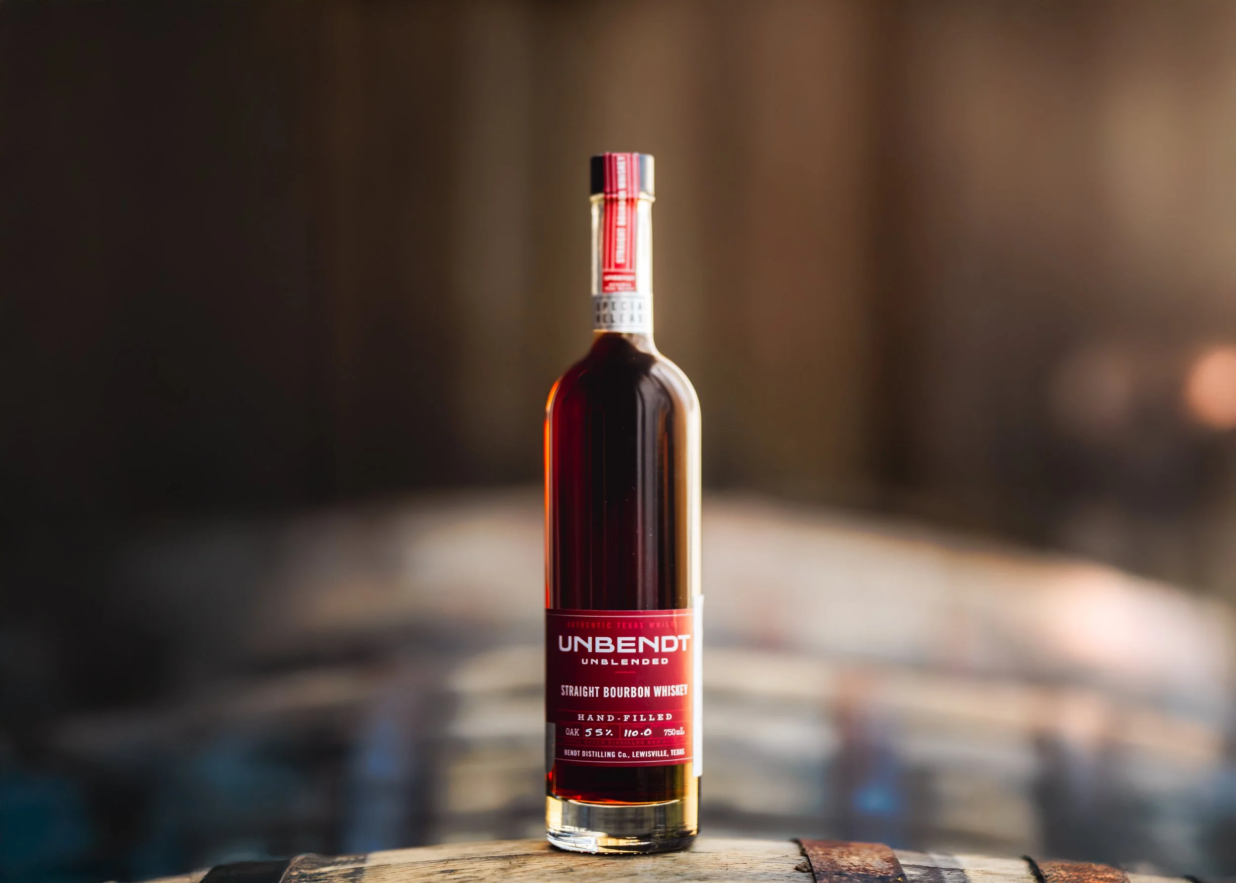 A bottle of Unbend Straight Bourbon Whiskey with a red label, standing on a wooden surface with blurred background.