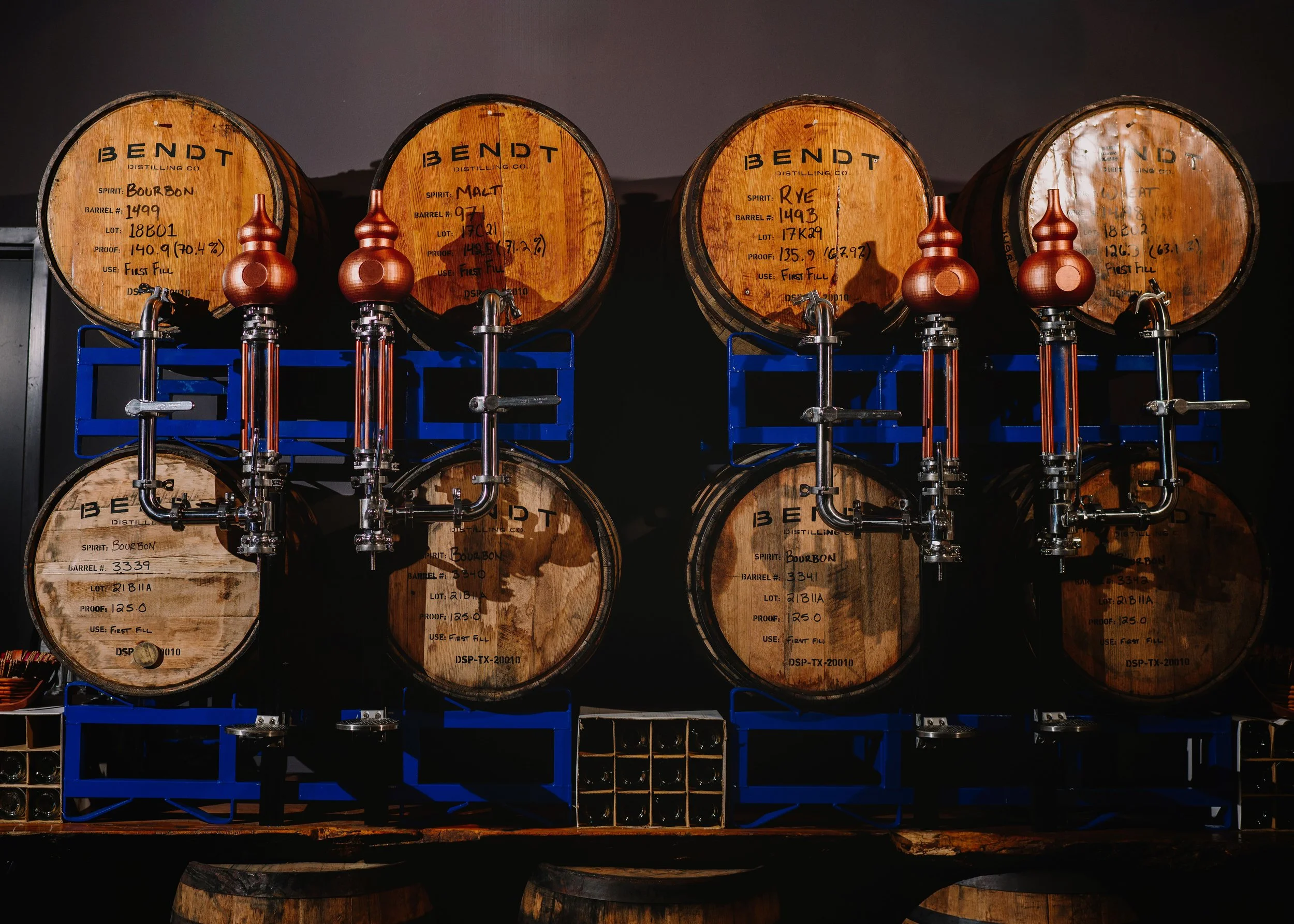 Four wooden barrels of bourbon whiskey on a blue metal rack, with spigots, against a dark background.