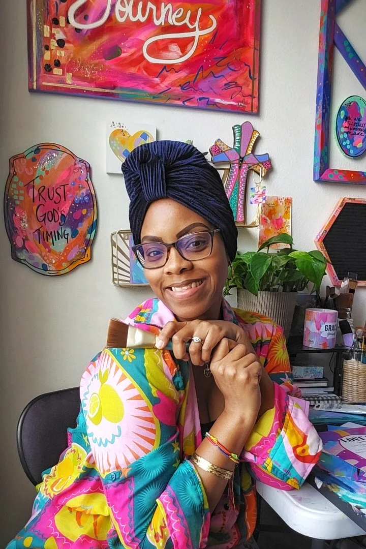 Desirae Ofori sitting in her art studio surrounded by colorful artwork. She is smiling, wearing a colorful blouse, a navy blue velvet turban and holding paint brushes.