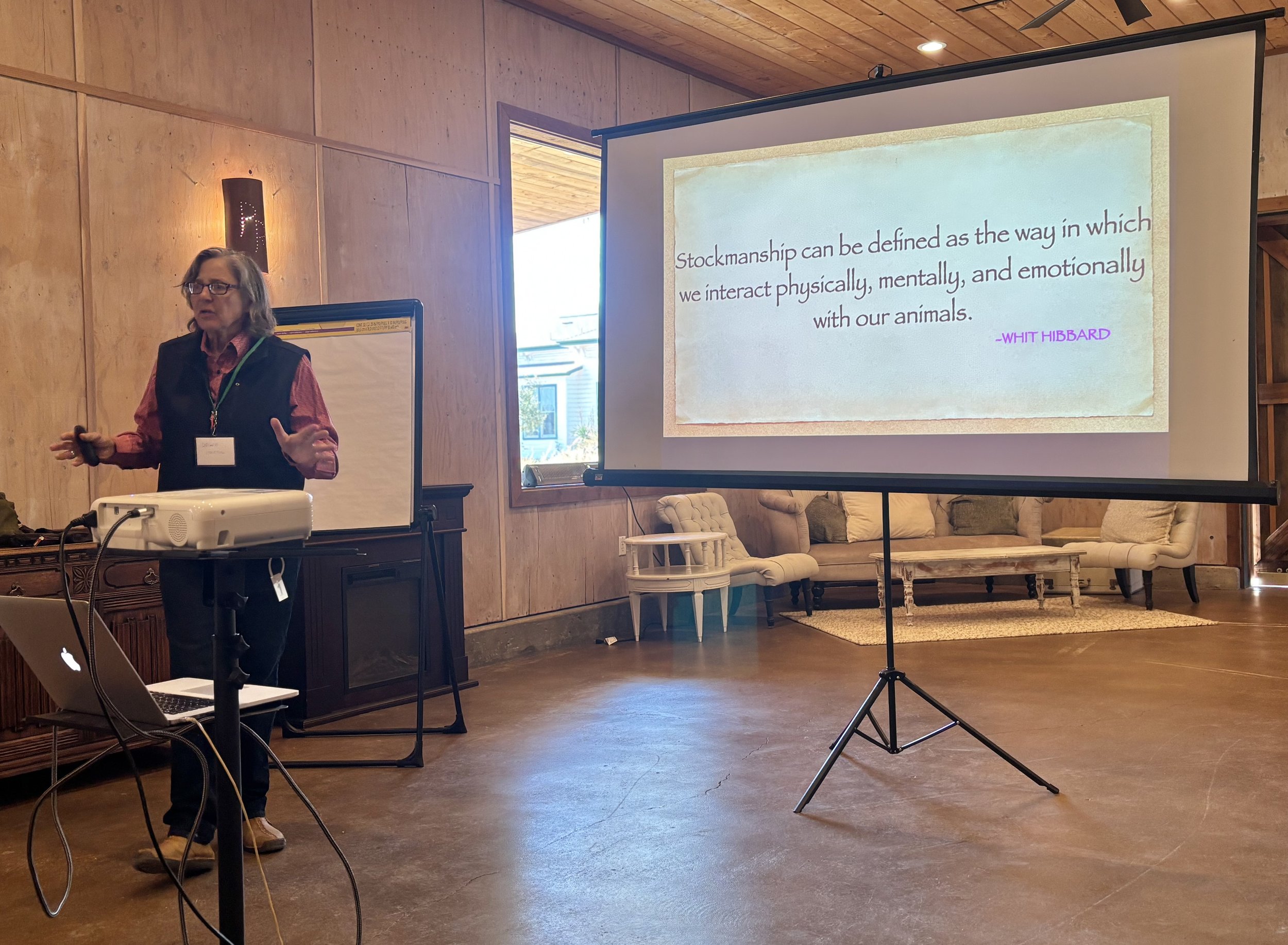 A woman giving a presentation on a stage with a projector and a large screen. There are chairs and a window in the background.
