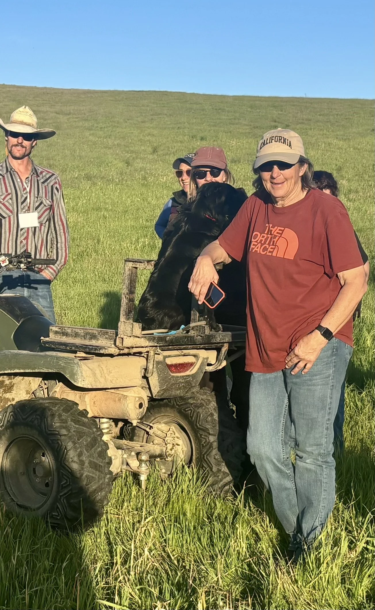 Group of people outdoors in grassy field, standing near a small ATV with a black dog sitting on the cargo bed.