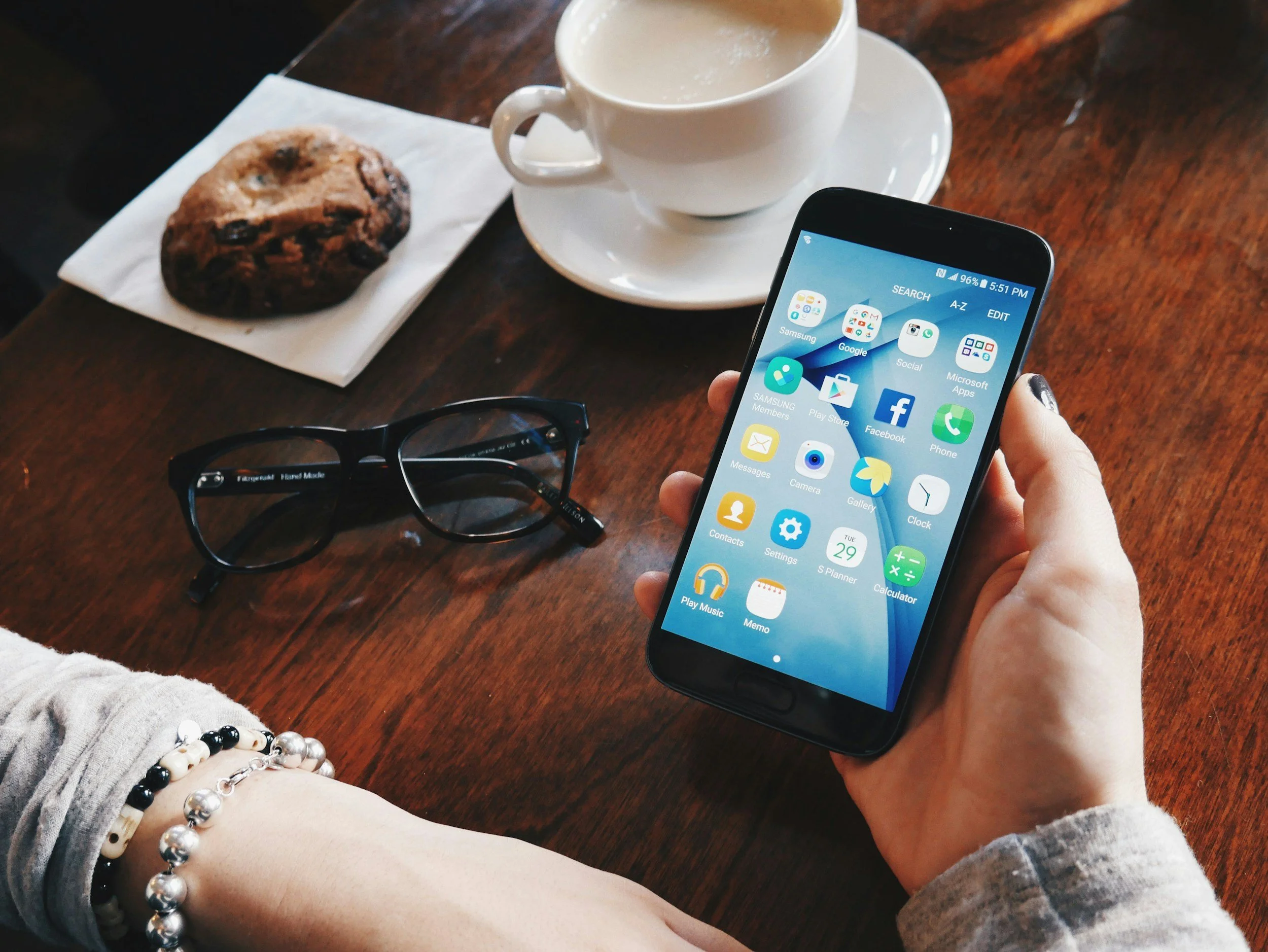 A person holding a smartphone displaying apps, sitting at a wooden table with a cup of coffee, a chocolate chip muffin, black glasses, and a beaded bracelet.