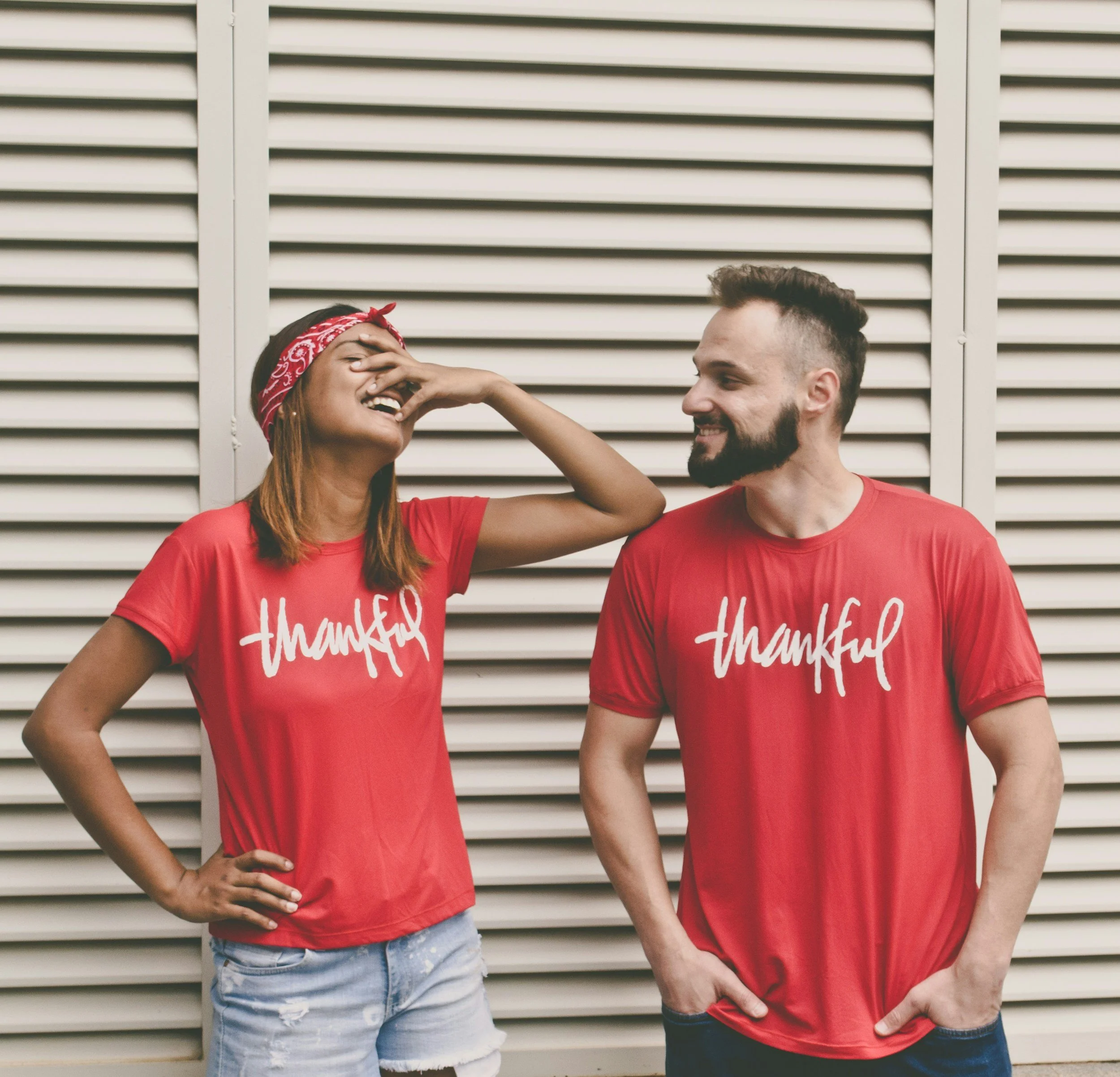 Two friends, a woman and a man, wearing red t-shirts with the word 'thankful' written on them, standing against a beige louvered wall. The woman is laughing and playfully pinching the man's nose while the man smiles at her.