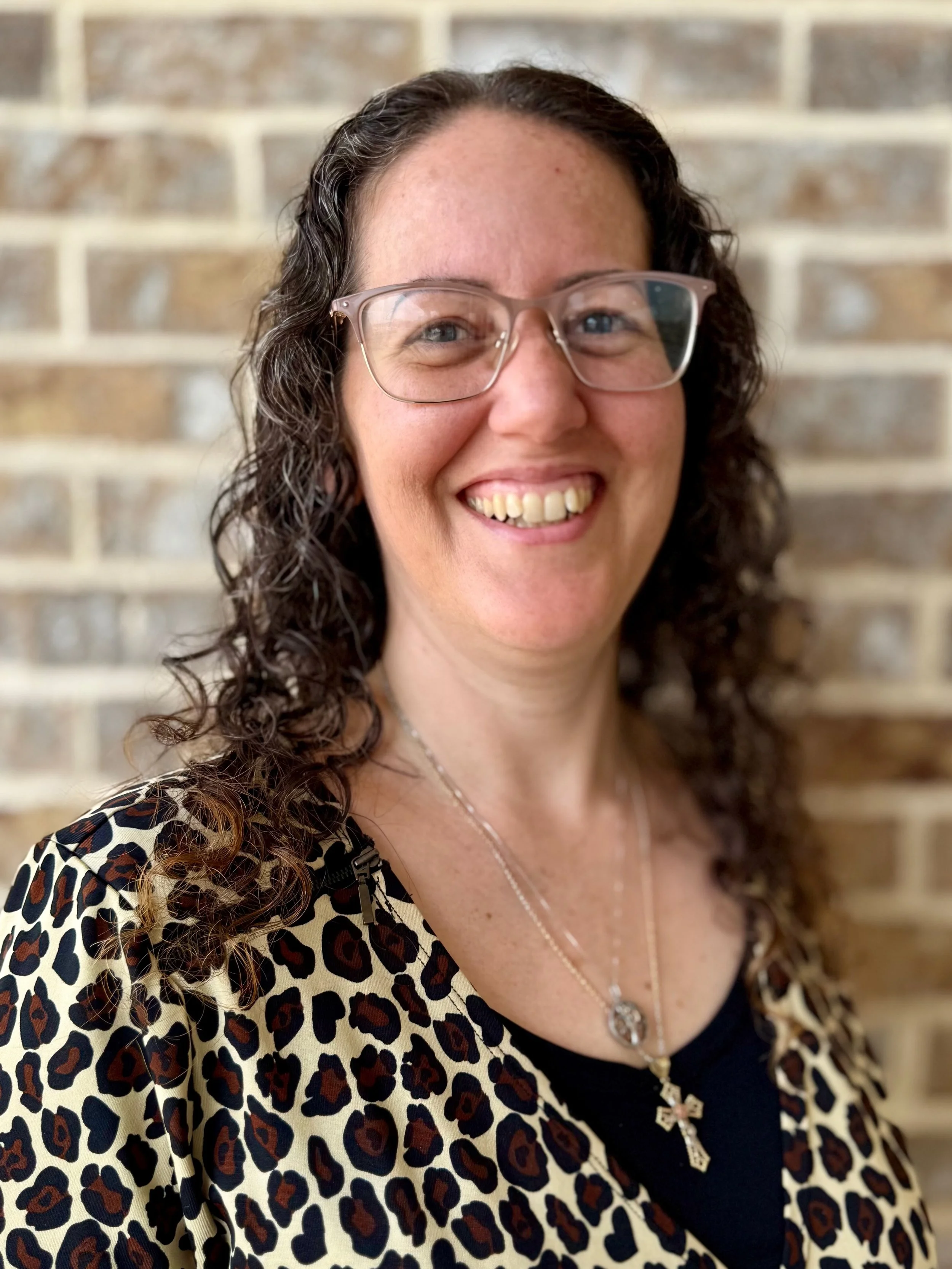 A woman with curly brown hair, glasses, and a cheerful smile, wearing a leopard print top, a black shirt, and a silver cross necklace, standing in front of a brick wall.