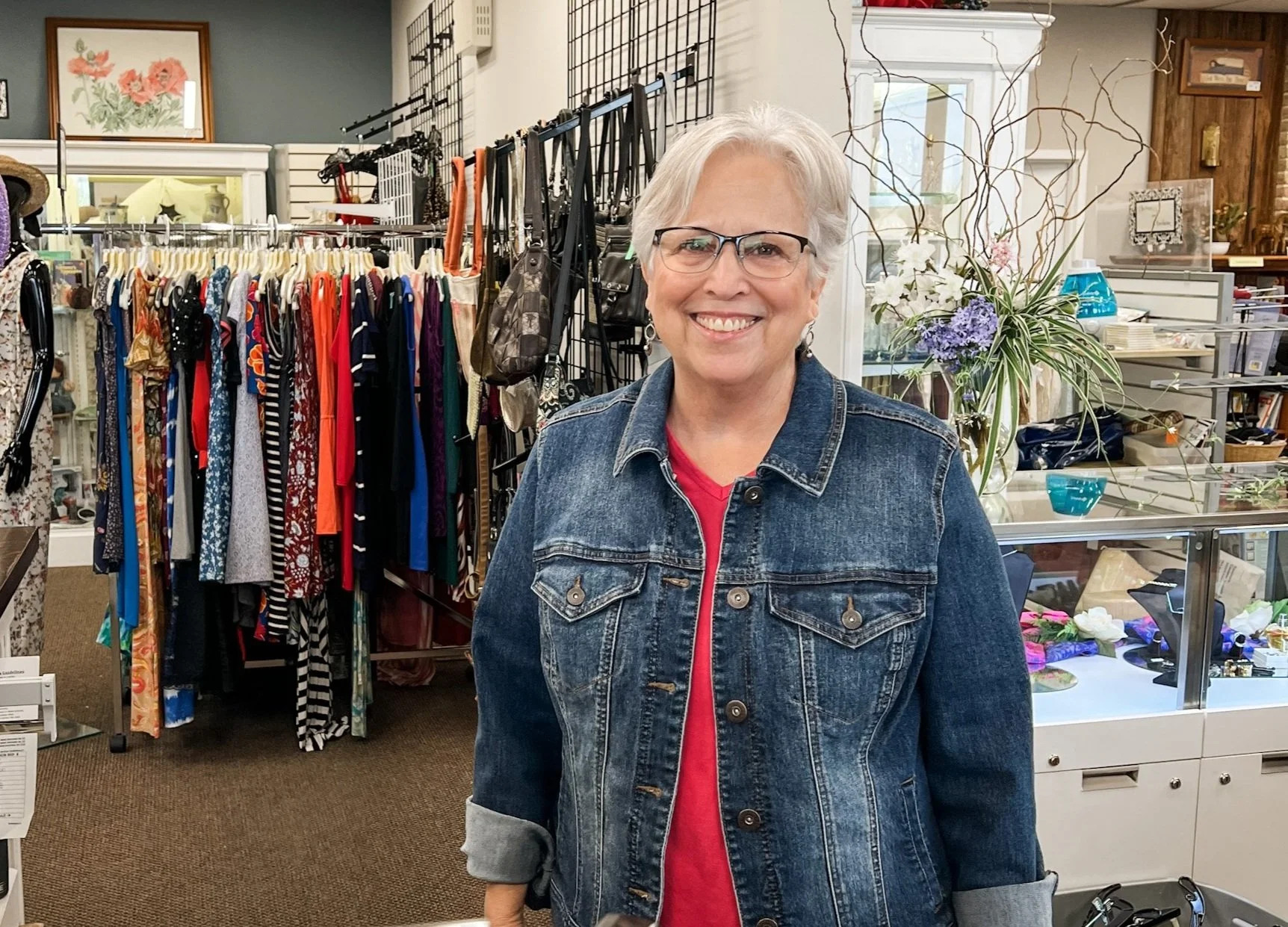 A smiling woman with short gray hair and glasses stands inside a thrift store, wearing a denim jacket over a red shirt. Behind her are clothing racks with various garments and display cases with jewelry and decorative items.