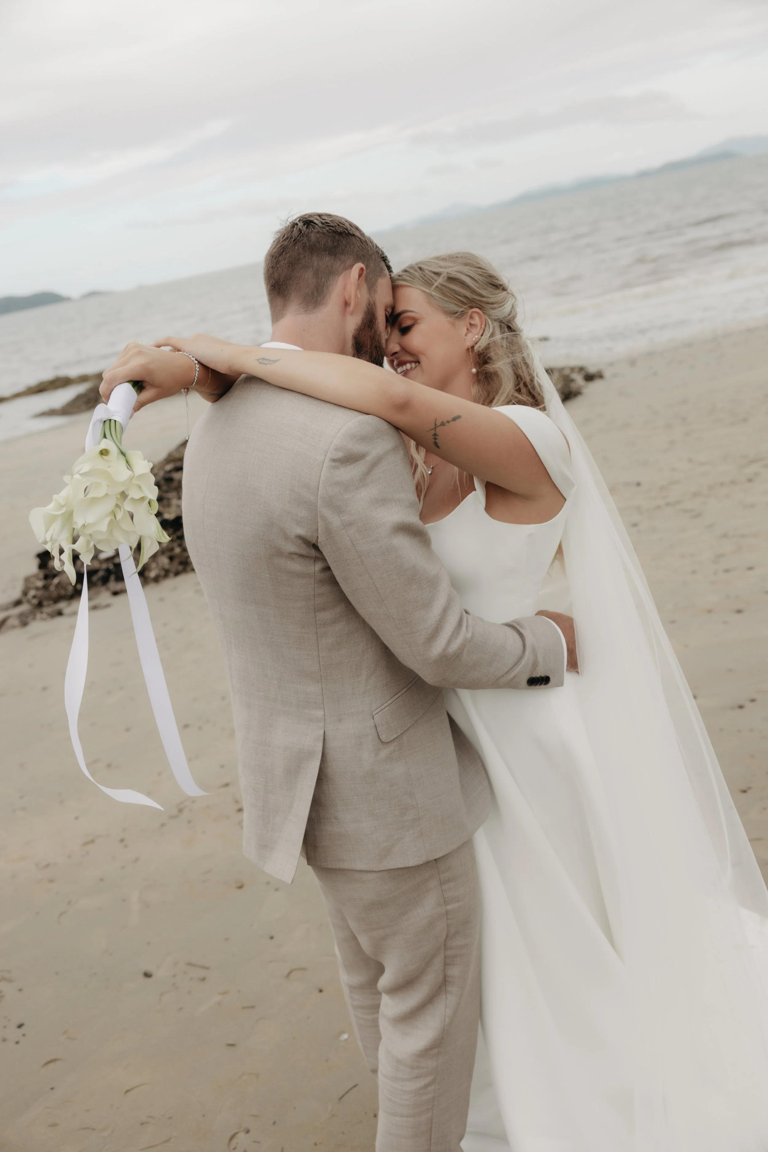 A couple dressed in wedding attire embracing on a beach with the ocean and cloudy sky in the background.