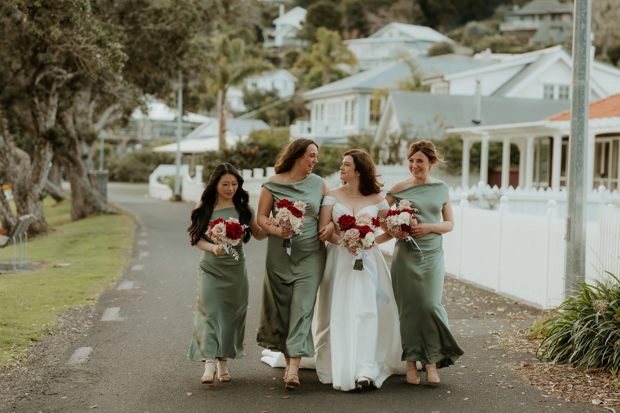 A bride in a white wedding dress walking with four women in matching green dresses on a residential street, holding bouquets of flowers, smiling and talking.