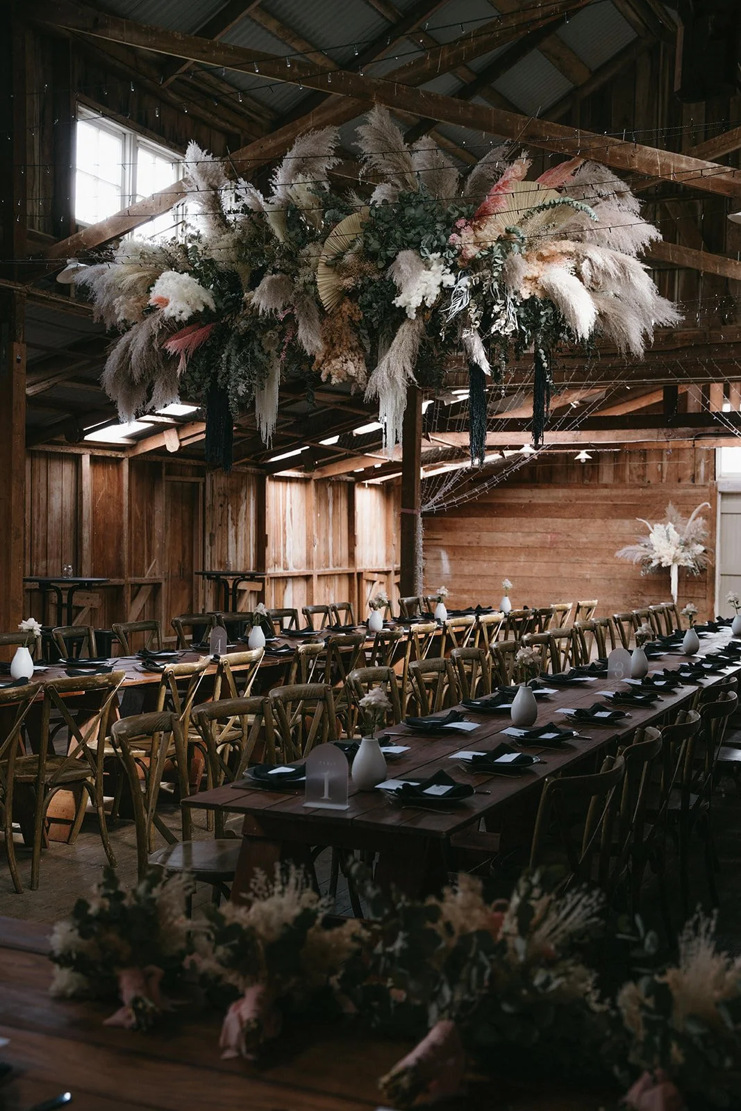 Long wooden banquet tables set with black napkins, white plates, and white vases with flowers inside. Overhead, a large floral arrangement with pampas grass and greenery hangs from the ceiling of a rustic barn, which has wooden walls and beams.