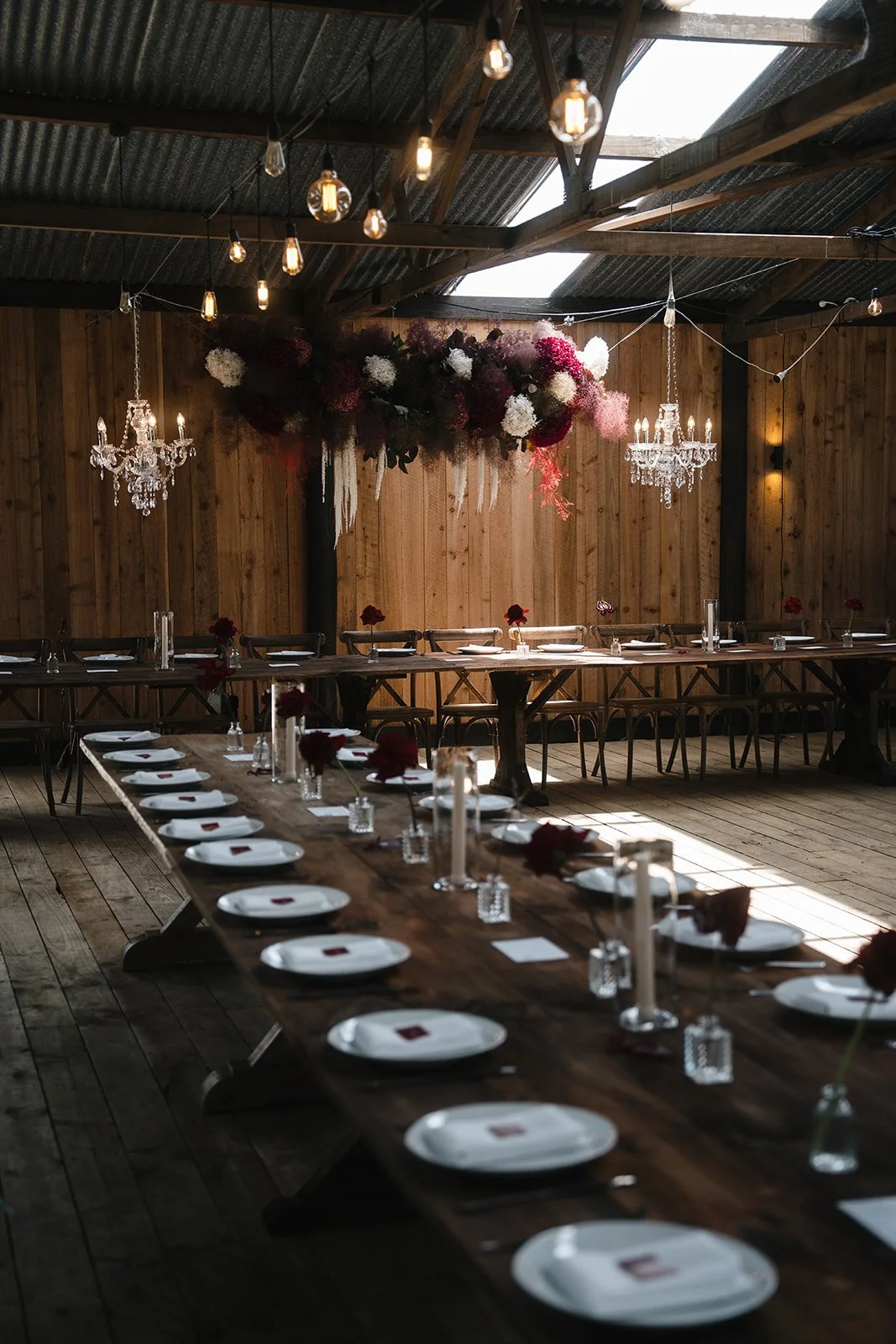 Rustic event space with long wooden tables set for a party, decorated with candles and red flowers, and a floral centerpiece with hanging chandeliers and string lights overhead.
