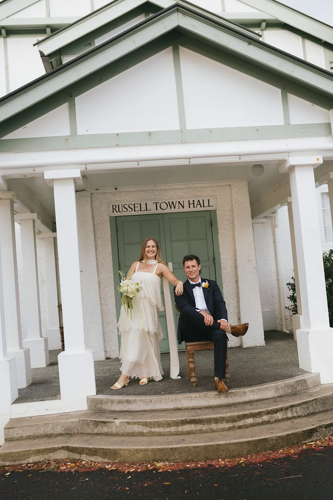 A newlywed couple dressed in wedding attire standing outside Russell Town Hall. The bride is holding a bouquet and resting her arm on the groom's shoulder, who is seated on a wooden chair with a pipe in his hand. The building is white with green accents, and the couple is smiling.