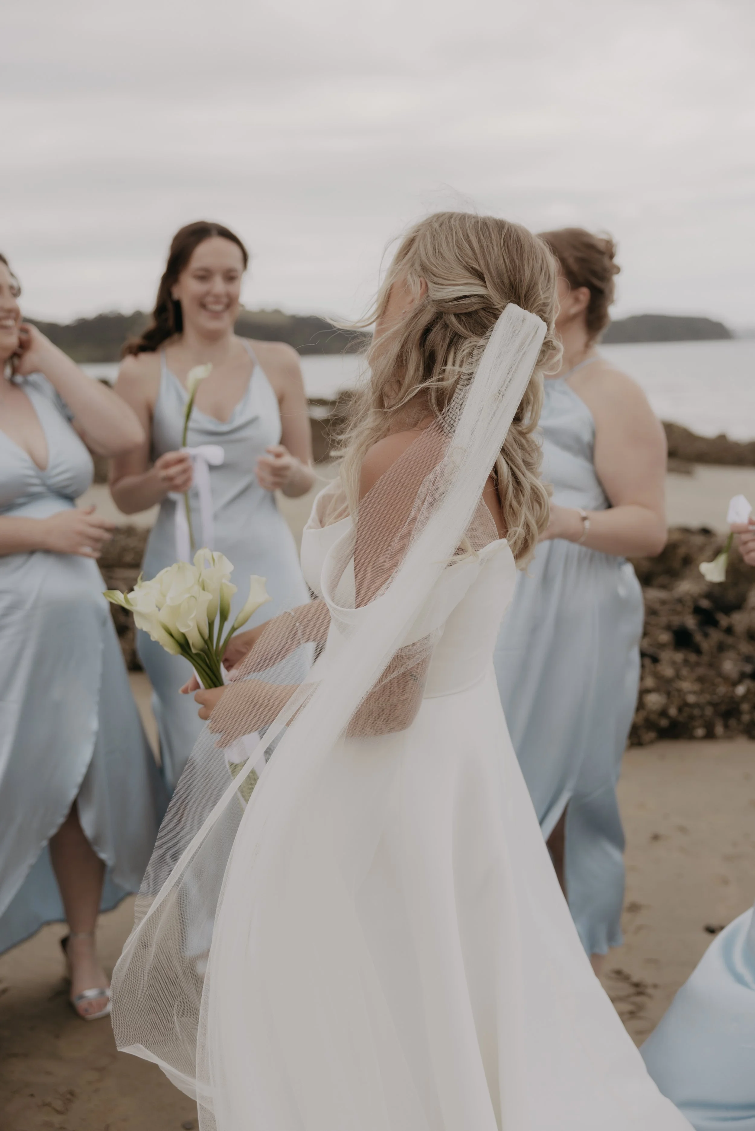 A bride in a white wedding dress holding a bouquet of white calla lilies, talking with bridesmaids on a beach, with rocky shoreline and ocean in the background.