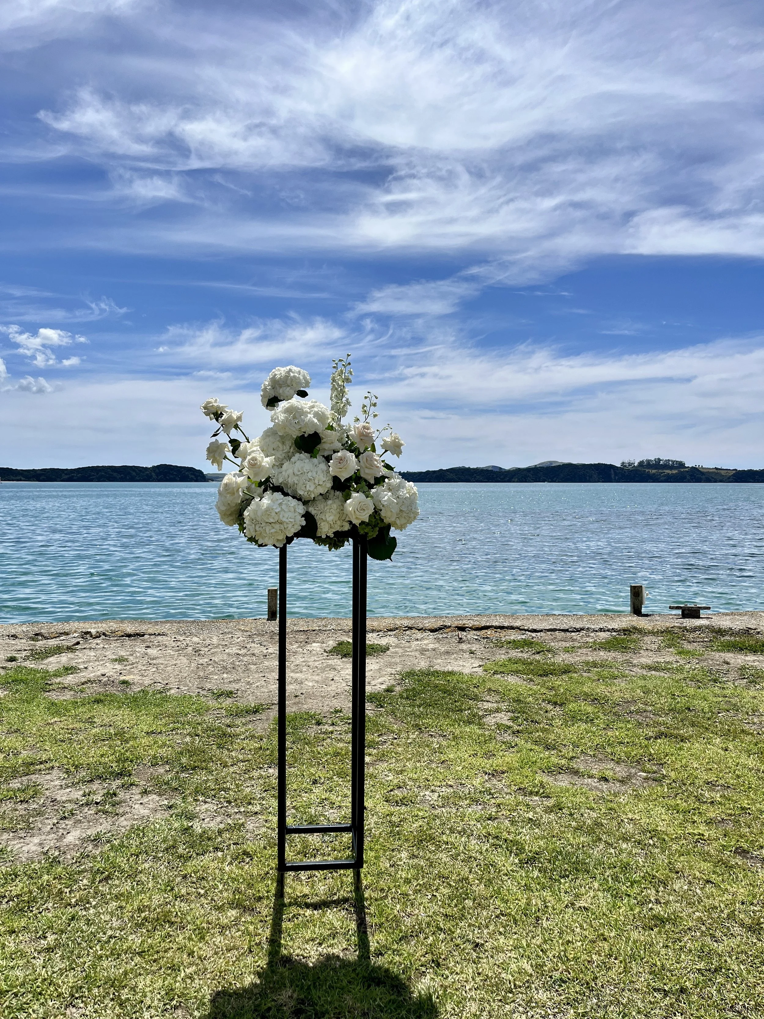 A floral arrangement consisting of white hydrangeas, roses, and other white flowers is placed on a black stand near a waterfront with islands in the distance, under a partly cloudy sky.