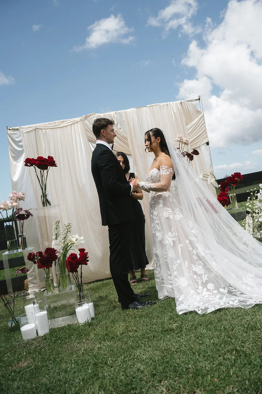 A bride and groom exchanging vows outdoors under a fabric backdrop with flowers, on a grassy field, with a partly cloudy sky.