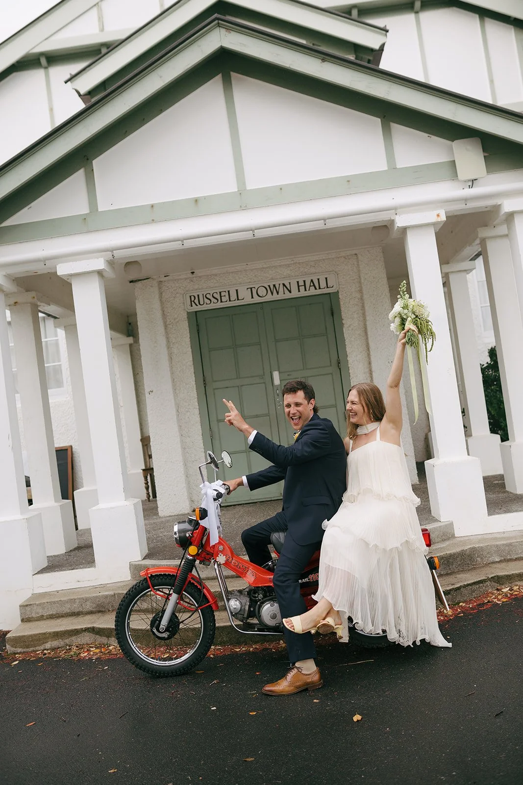 A newlywed couple celebrating on a red motorbike in front of Russell Town Hall. The groom is dressed in a suit, and the bride in a white dress holding a bouquet with her arm raised. Both are smiling and appear happy.