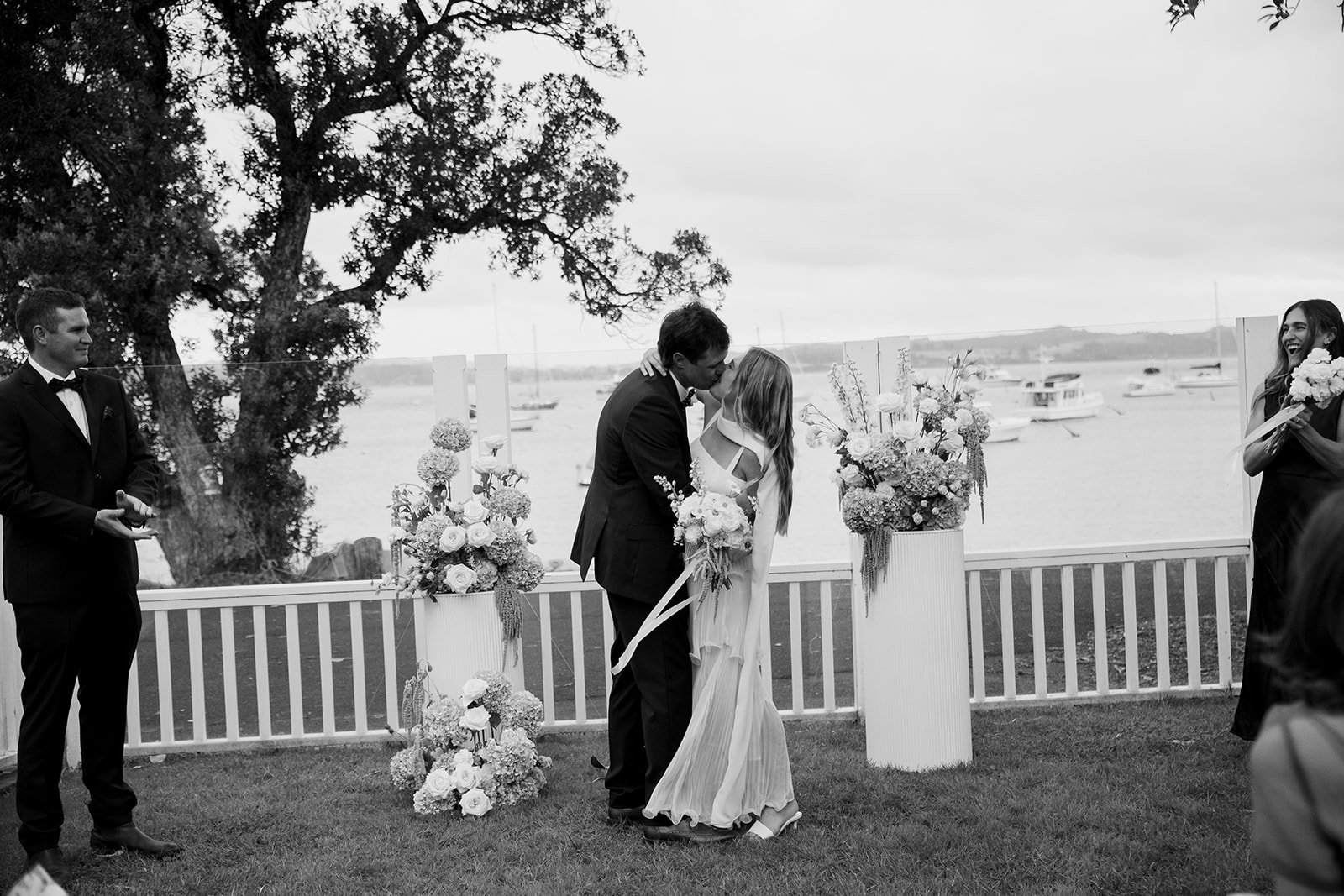 A wedding ceremony by the water with a couple kissing under flower arrangements, surrounded by friends, sailboats in the background, and a large tree.