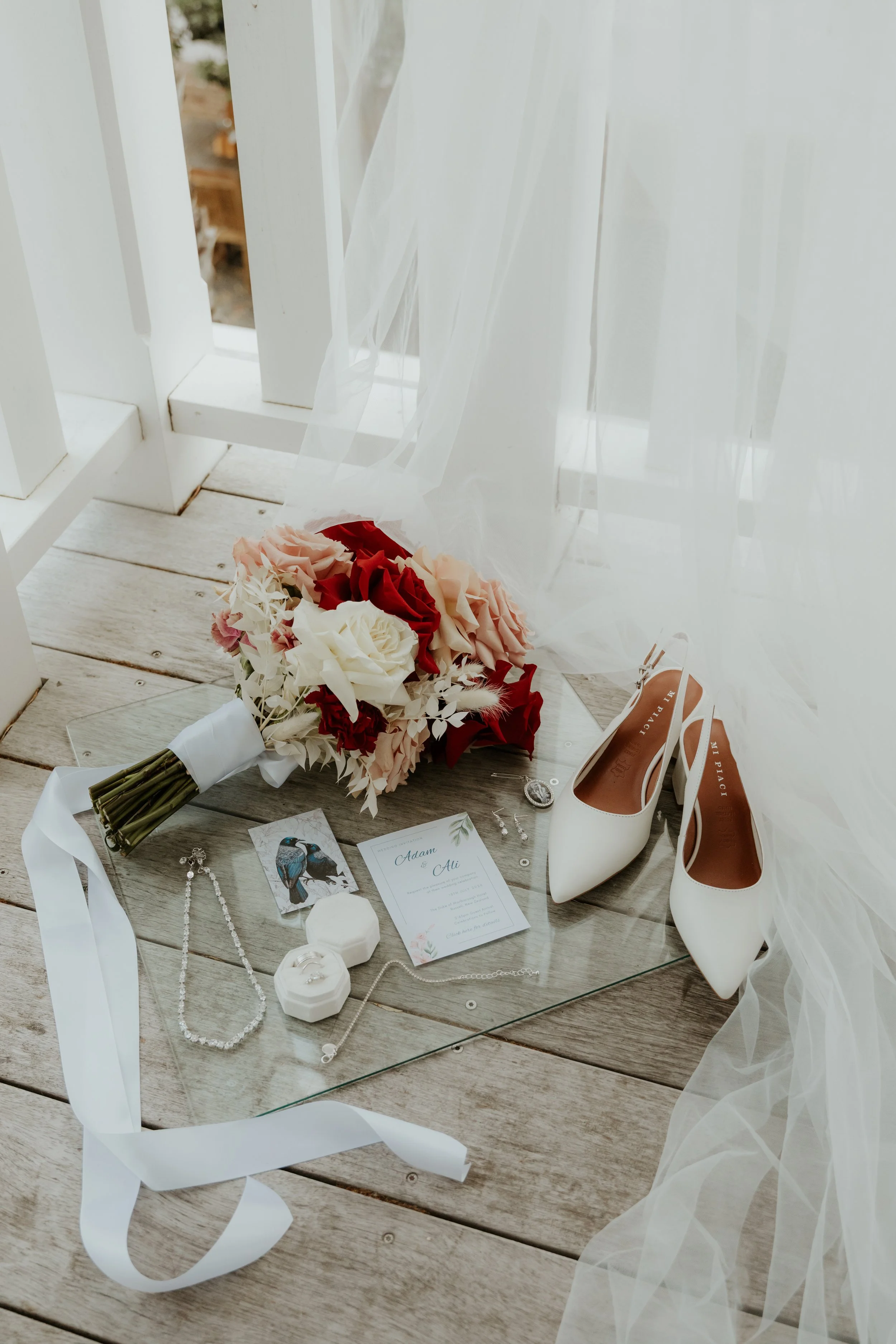A wedding bouquet with pink, red, and cream flowers, a pair of white high-heeled shoes, a wedding invitation, jewelry, and other small accessories on a glass table near a window with sheer white curtains.