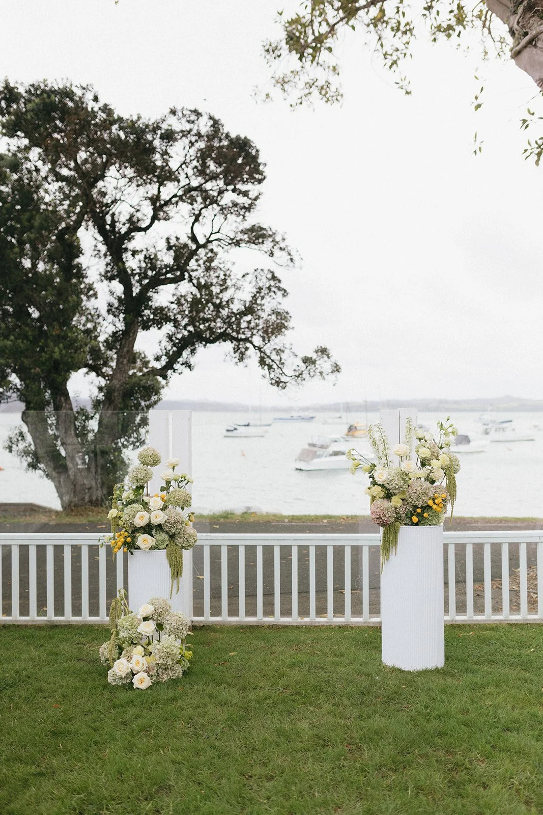Festival or event setup with two tall white vases filled with white and pale purple flowers, set outdoors near water with boats in the background, on a grassy area behind a white railing.