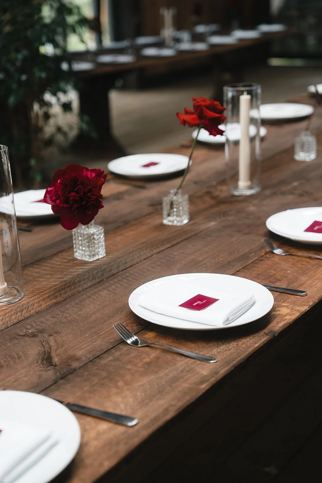 A rustic wooden table set for a meal with white plates, silver forks, white napkins with red place cards, and a row of small glass vases with red and orange flowers. There are also tall glass candle holders with white candles.