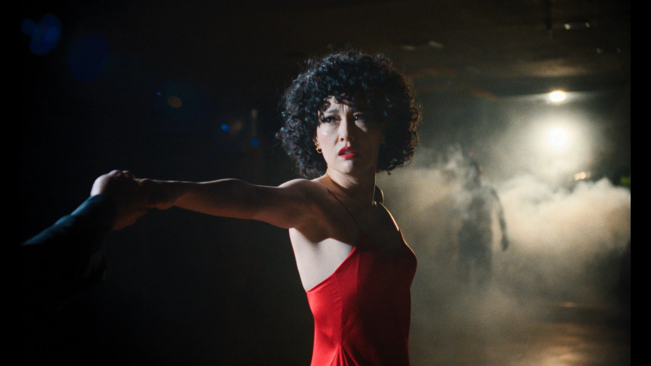 Rinko Kikuchi as Haru in a red slip dress, arm extended in a sharp dance pose, silhouetted figures in haze behind her under warm stage light.