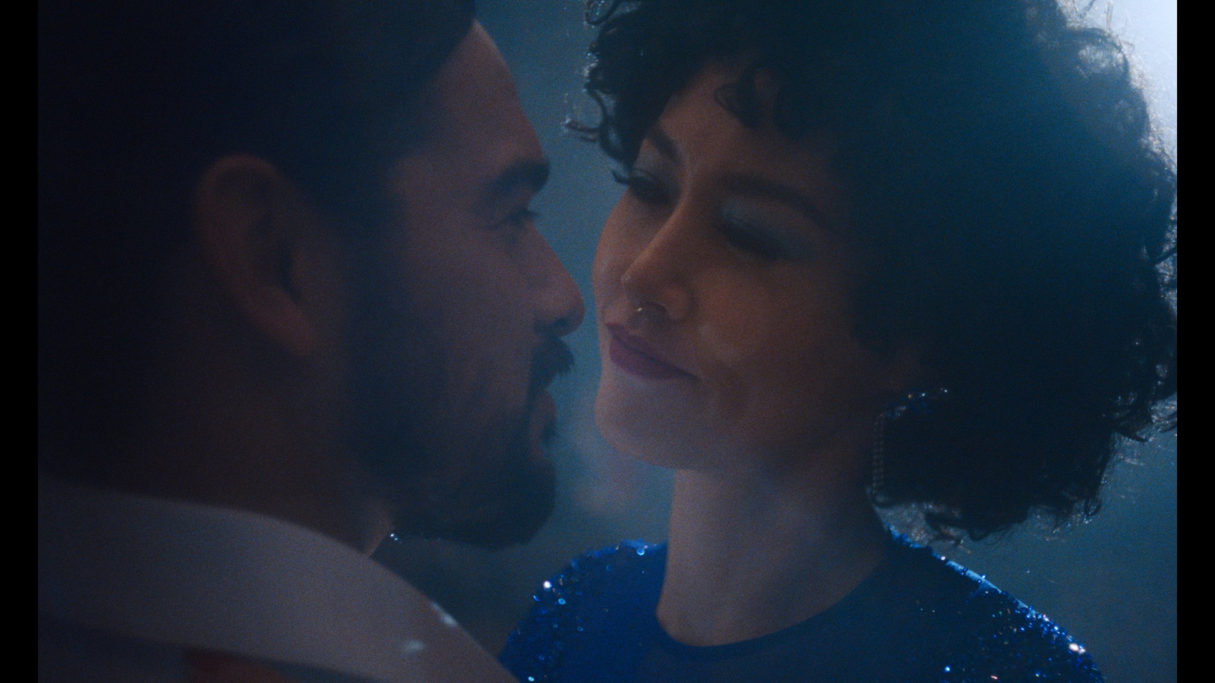 Rinko Kikuchi as Haru and Alejandro Edda as Luis in close-up profile, faces close together in an intimate moment, softly lit in blue tones.
