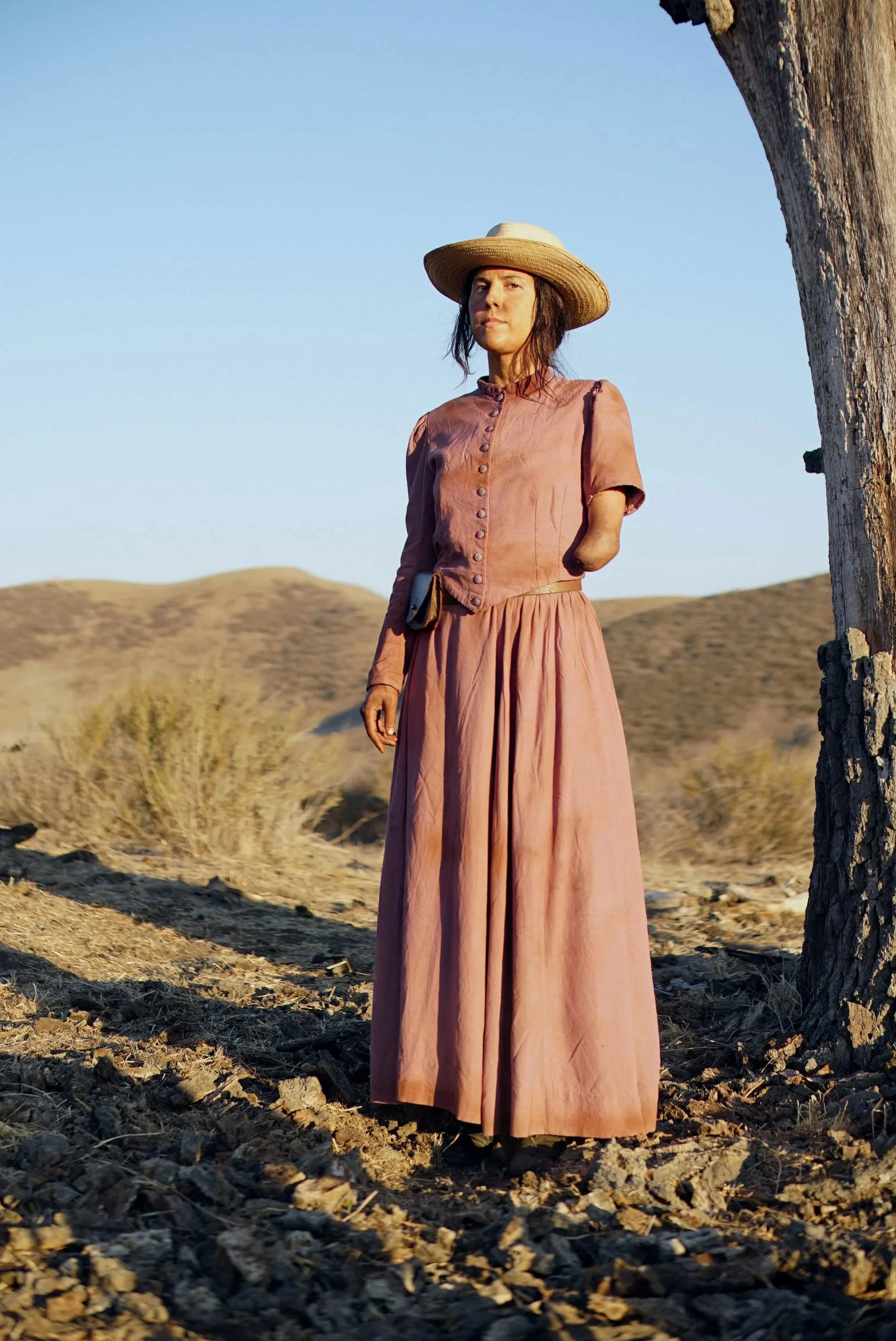 Angel Giuffria dressed in a pink vintage style dress and vest, wearing a wide-brimmed straw hat, standing outdoors in a desert landscape with sparse vegetation and hills in the background. Costume design by Keagan Roberson for 'To The Dust'.