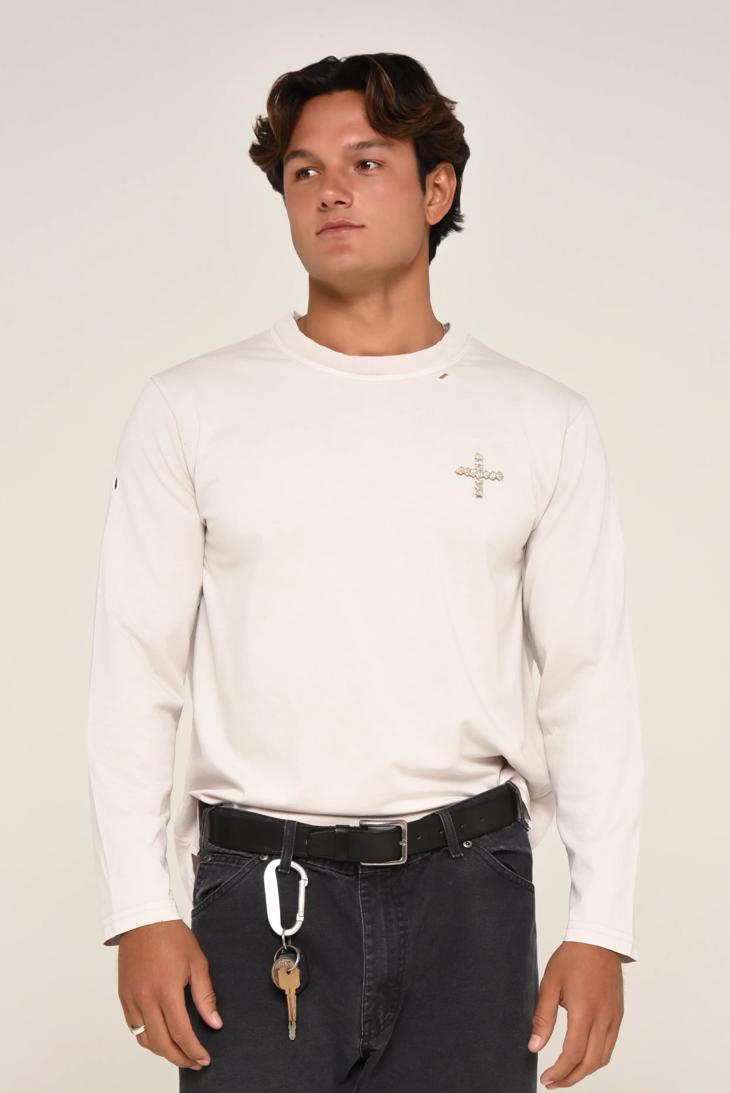 Model wearing a white long-sleeve shirt with a small cross emblem, styled with black jeans and a belt detail - designed in collaboration with Keagan Roberson.