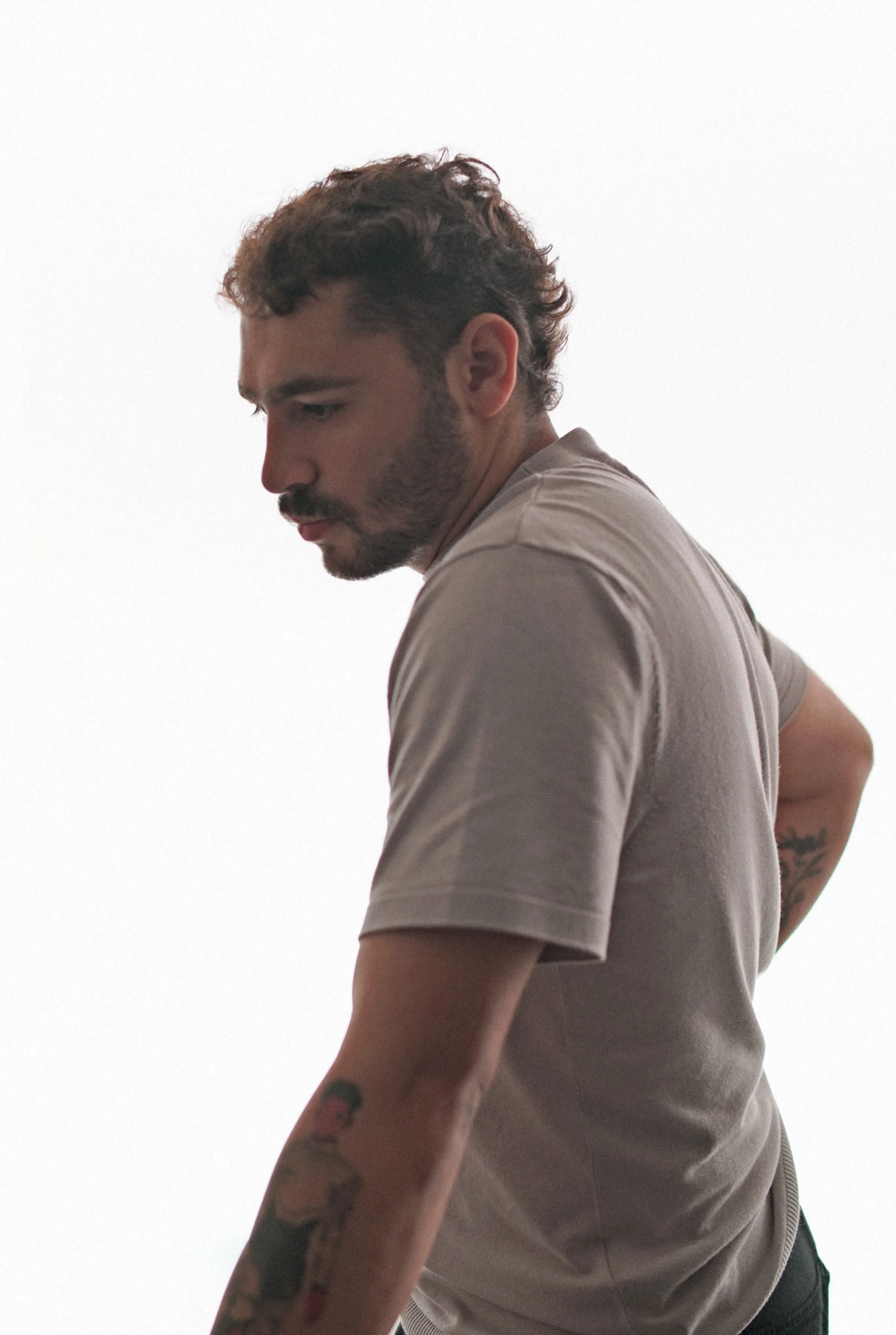 A young man with curly brown hair and tattoos on his arm, looking down and to the side, wearing a beige t-shirt against a plain white background.