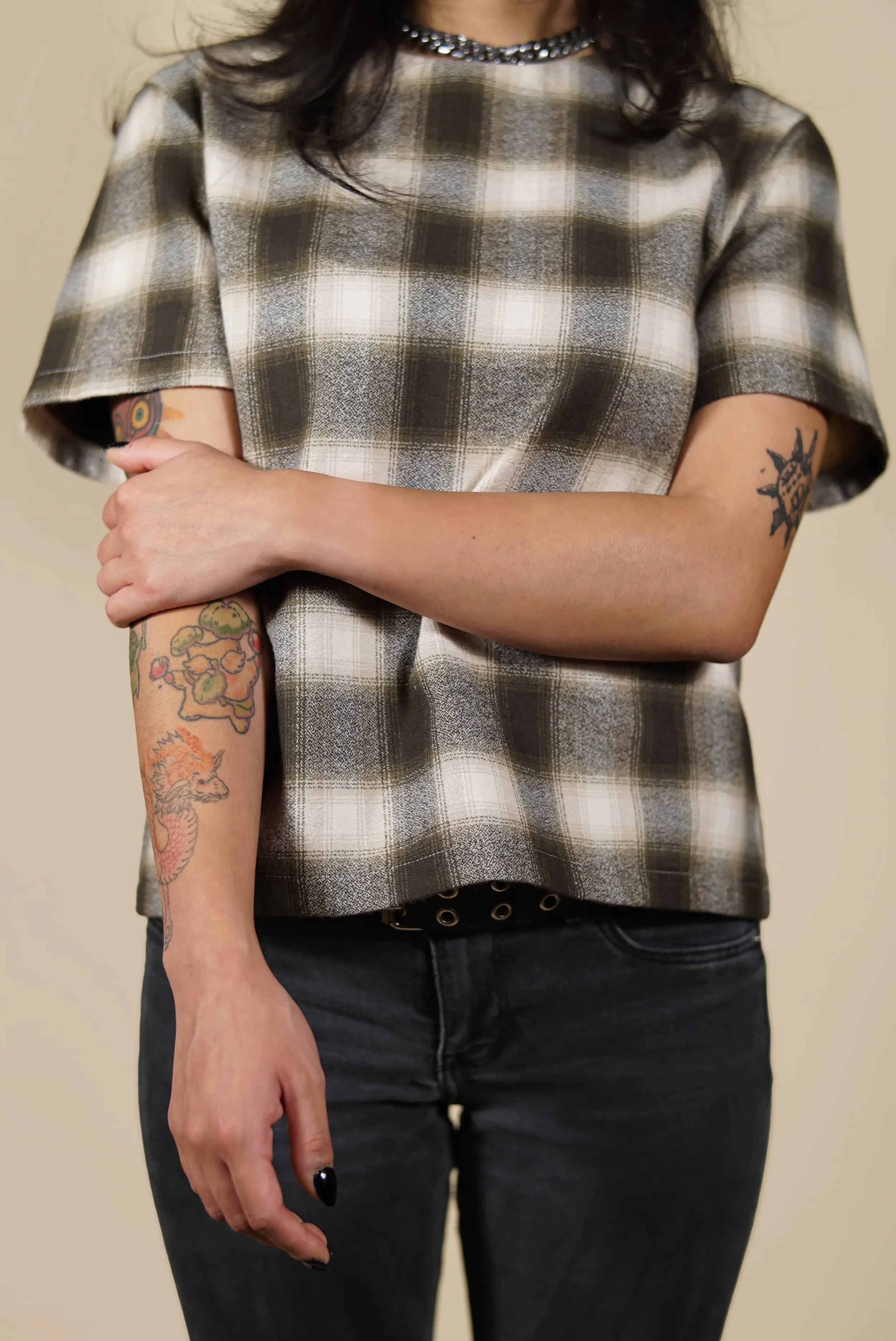 Person with tattoos on both arms, wearing a plaid short-sleeve shirt and black jeans, with their arms crossed.