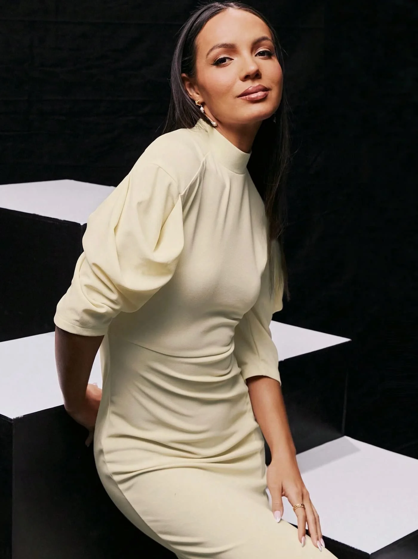 Model in a cream puff-sleeve dress from Keagan Roberson, seated on a geometric platform - elegant contrast of light and form.