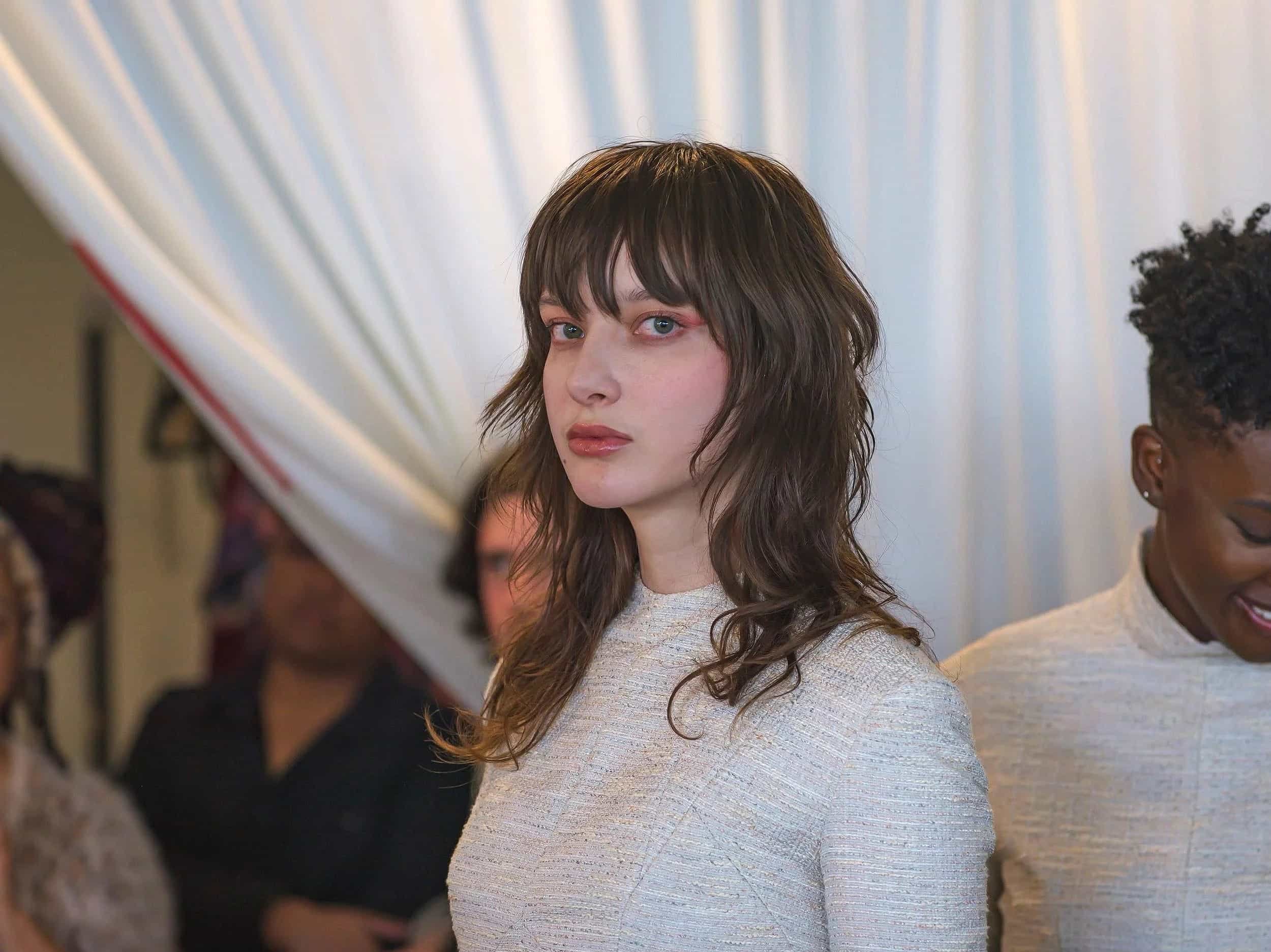 A woman with shoulder-length brown hair and bangs, wearing a white textured top, looking at the camera with a neutral expression. Other people are in the background at an indoor event.
