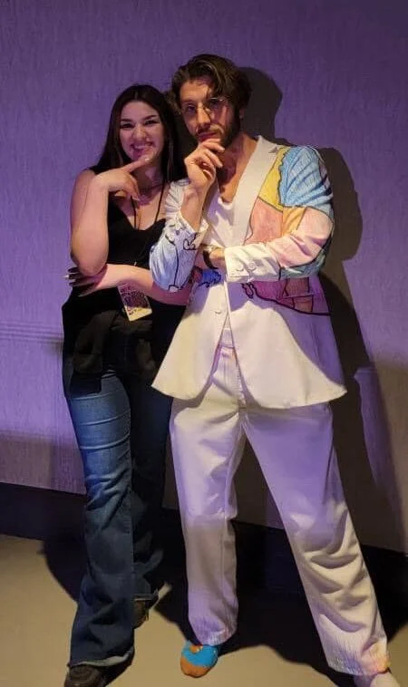 Olivia Bevacqua standing next to a man dressed in colorful, retro-style clothing, both posing with playful expressions against a plain purple wall.