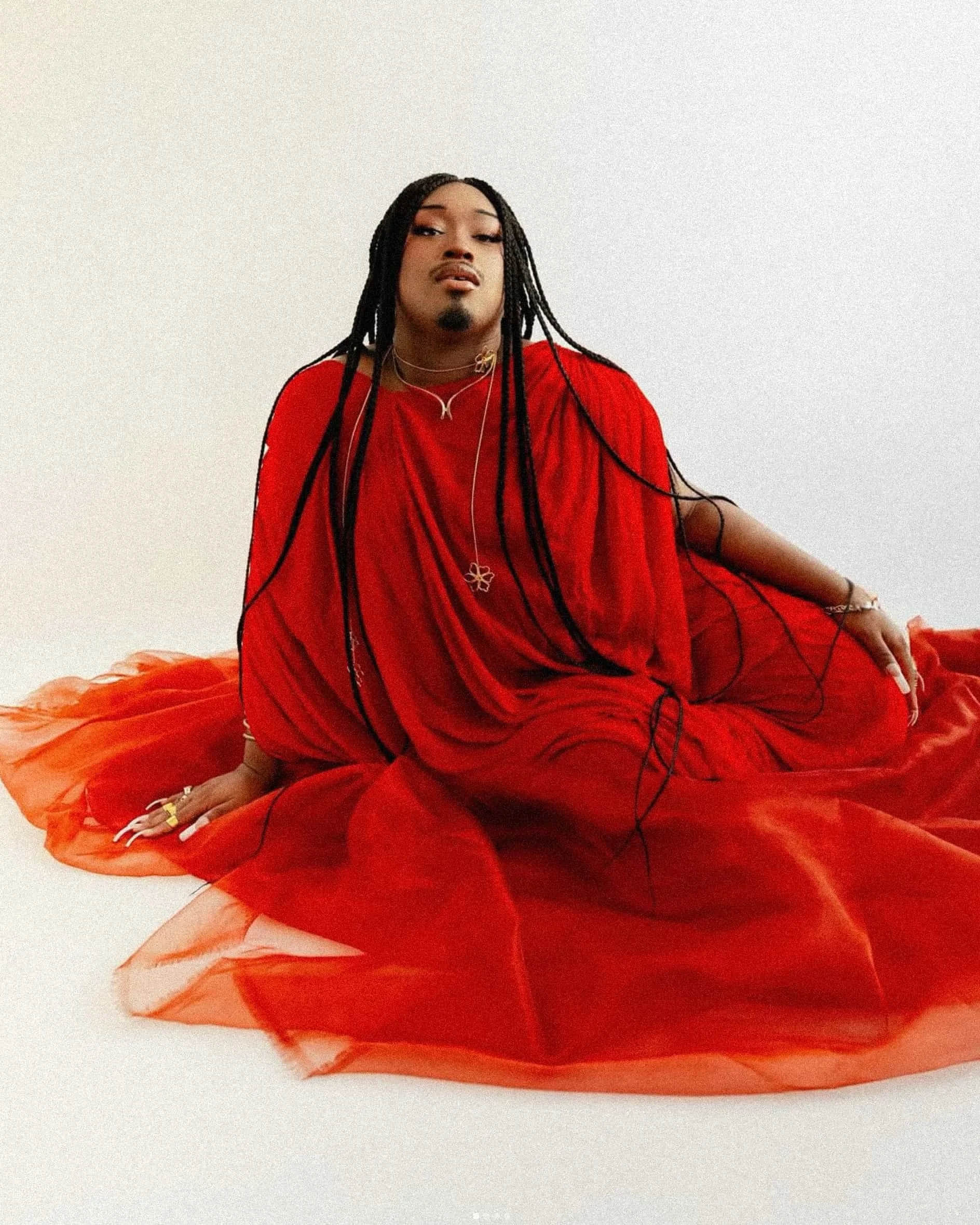 Cyncir "Foxxi" Pollard wearing a flowing red dress with layered jewelry, photographed against a white backdrop.