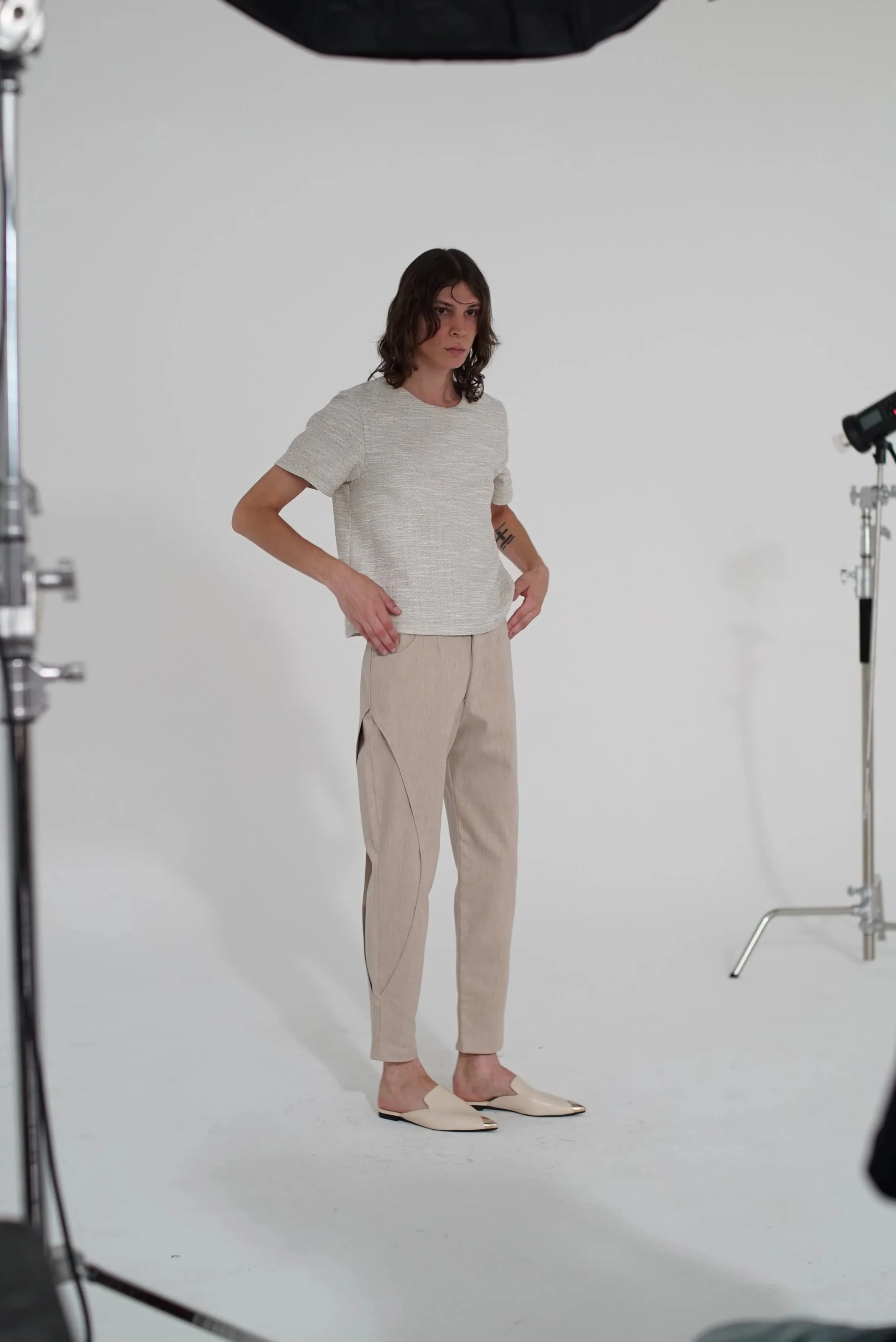 A woman standing in a photo studio, wearing beige pants, a light-colored short-sleeve shirt, and cream-colored slip-on shoes, with studio equipment visible around her.