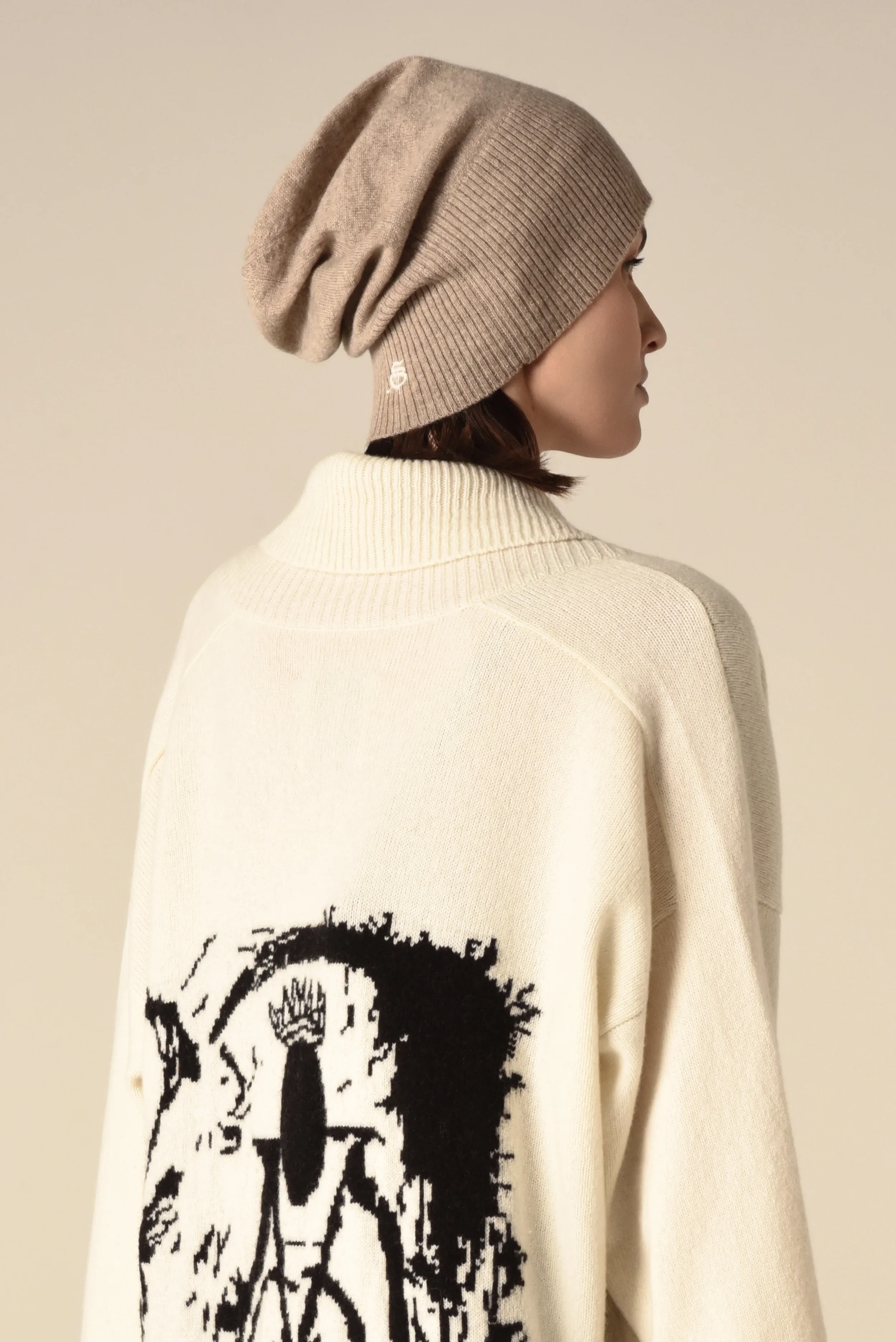 Side view of a model wearing a beige beanie and oversized cream sweater with black graphic print - designed in collaboration with Keagan Roberson.