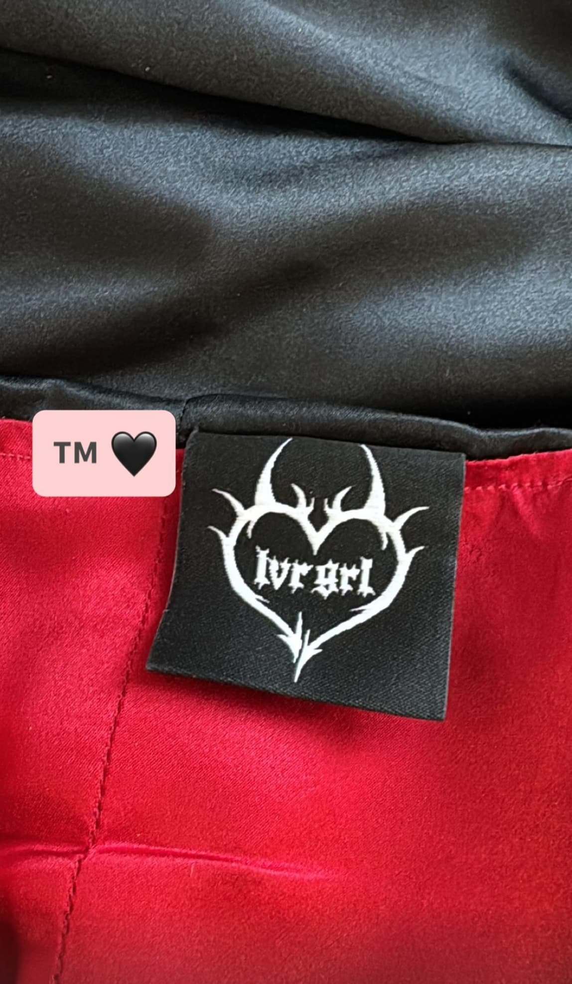 Close-up of a custom clothing label reading LVRGRL inside a black heart, sewn onto red and black fabric.