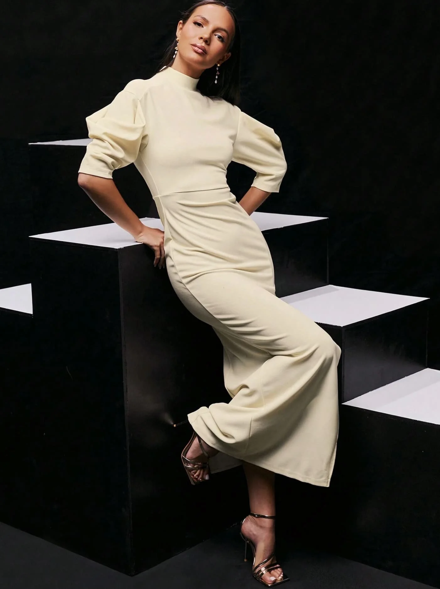 Model in a cream dress from Keagan Roberson, posing on black-and-white stairs. Sculptural composition blending movement and elegance.