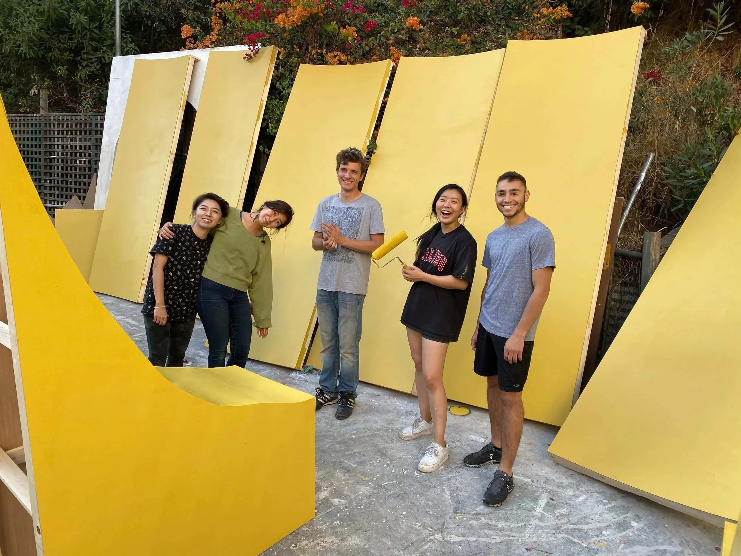 Keagan Roberson, Marc de Bertier, Rui Arichika and Mary Ma standing outdoors in front of large yellow wooden panels, smiling and enjoying a painting project; one person holding a yellow paint roller.