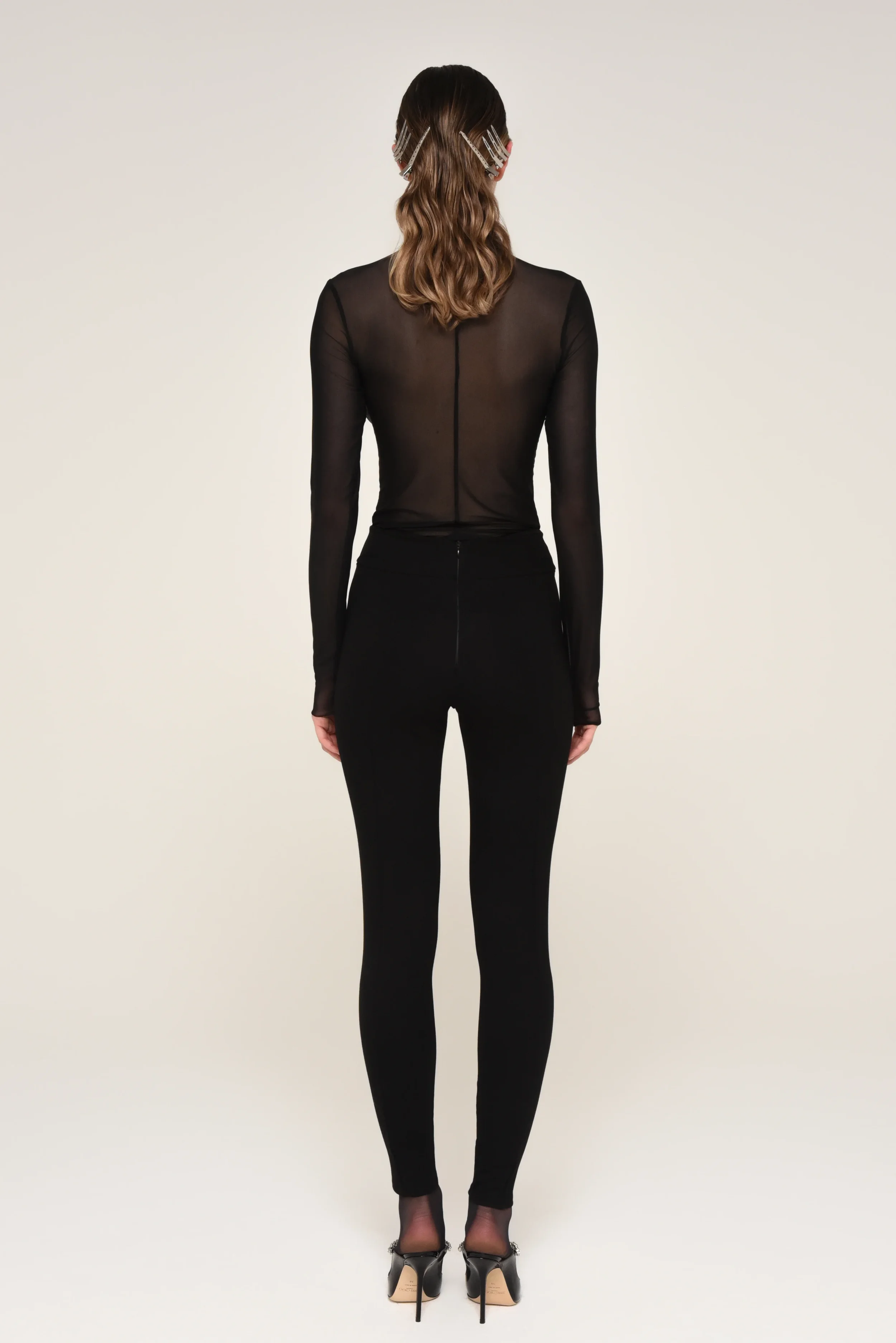 Back view of a model with wavy hair wearing a sheer black top and tailored black pants - designed in collaboration with Keagan Roberson.