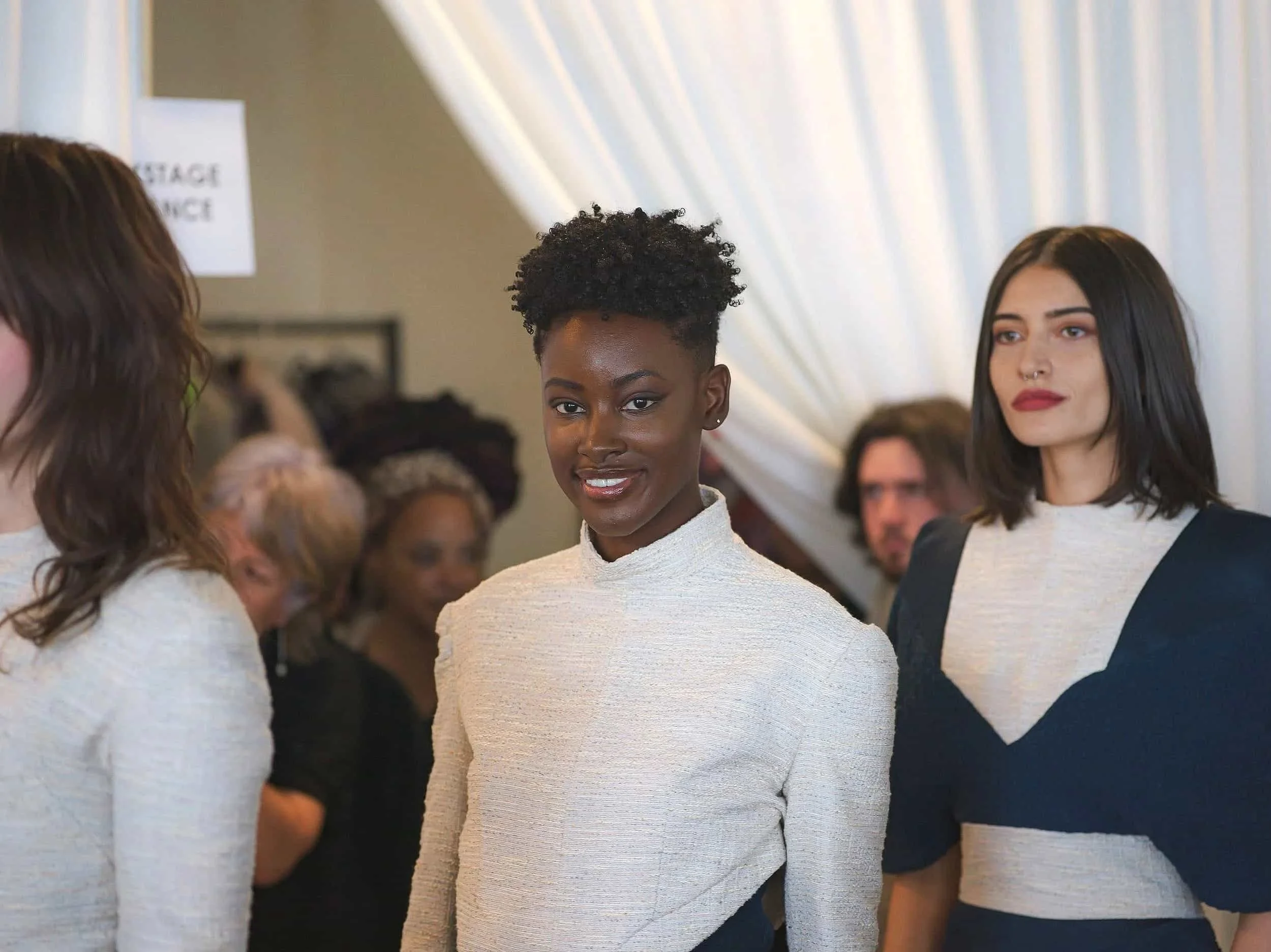 Group of diverse women in stylish clothing at a social event, with a woman smiling in the center and others chatting in the background.
