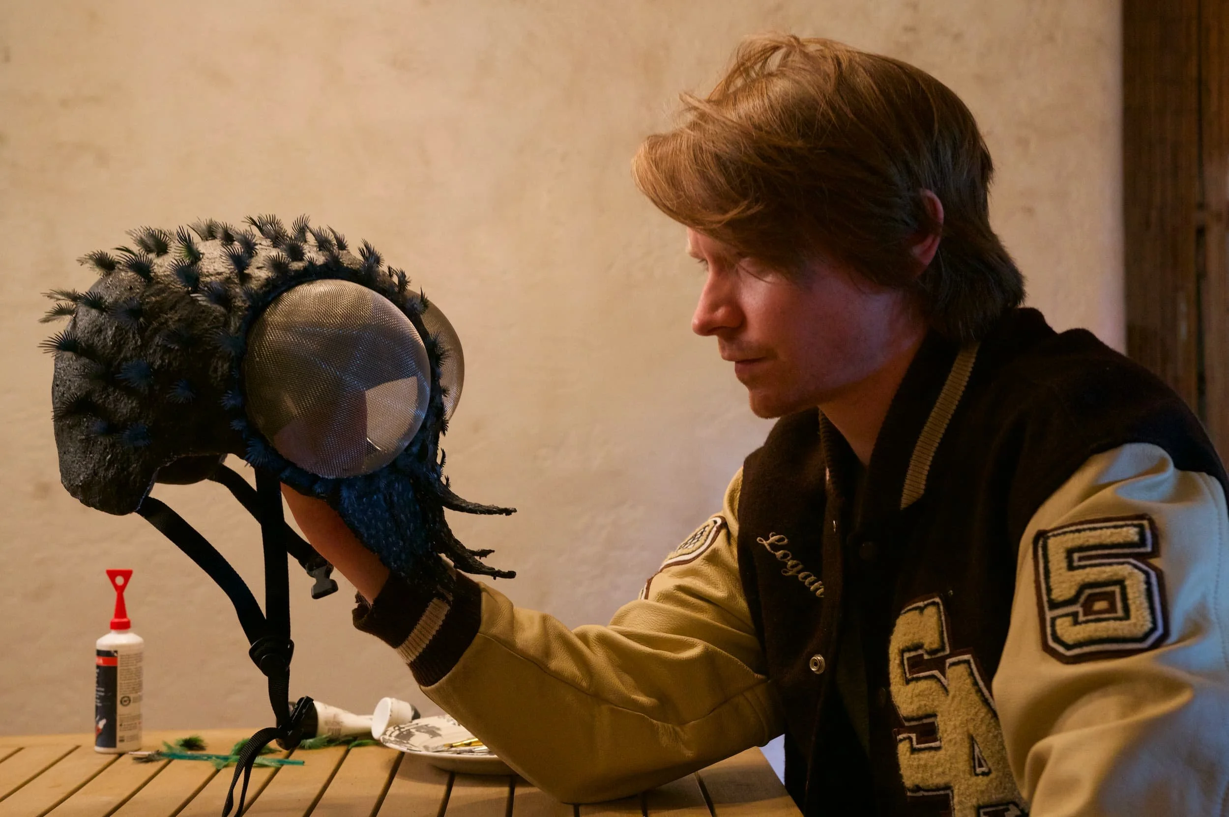Calum Worthy wearing a beige and black letterman jacket, holding a hand-crafted bug helmet with faux hair and eyes - costume piece designed by Keagan Roberson for Shedding Angels.