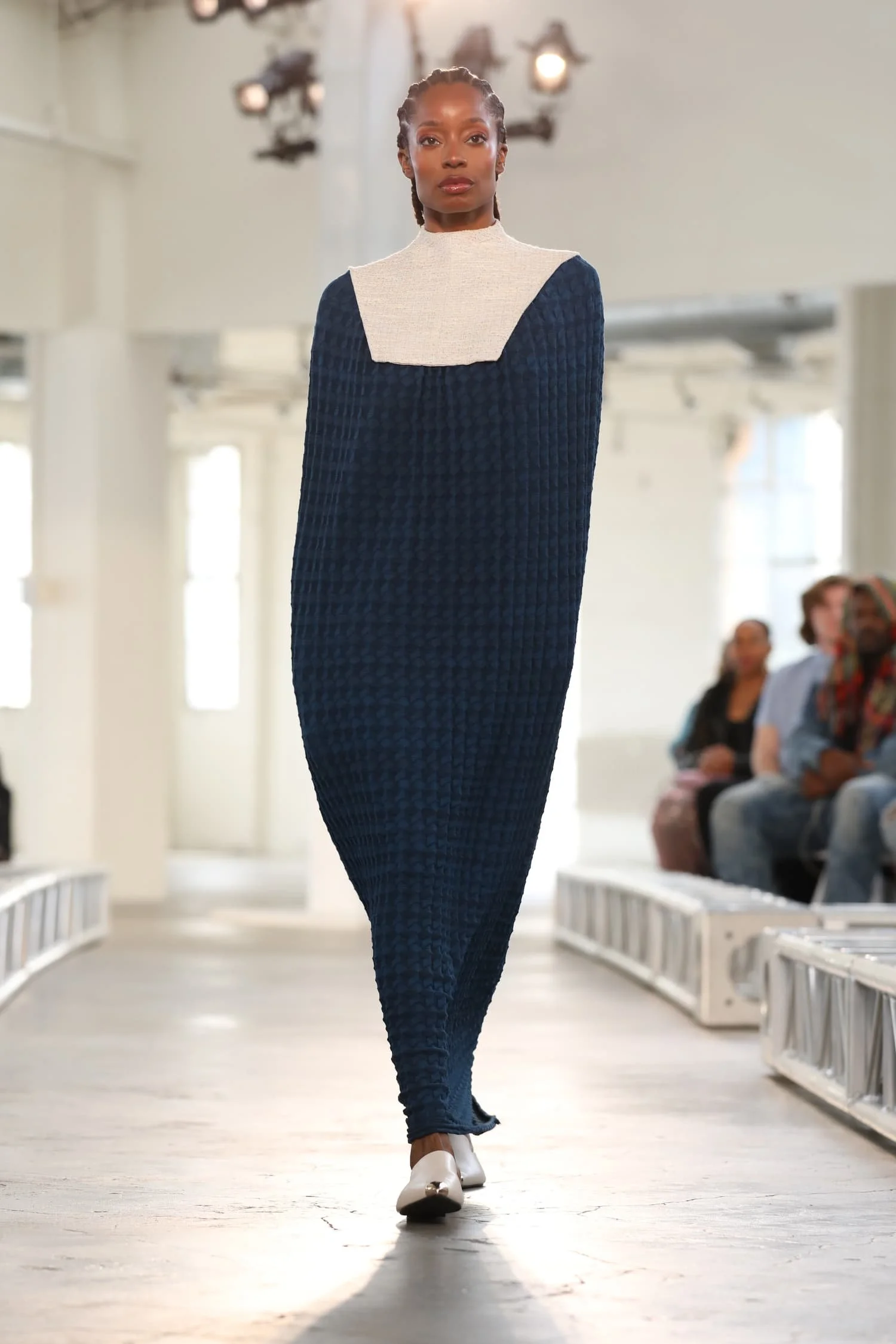 A model walking on a fashion runway wearing a long, textured blue dress with a white high-neck top underneath, and white shoes. Keagan Roberson collection.