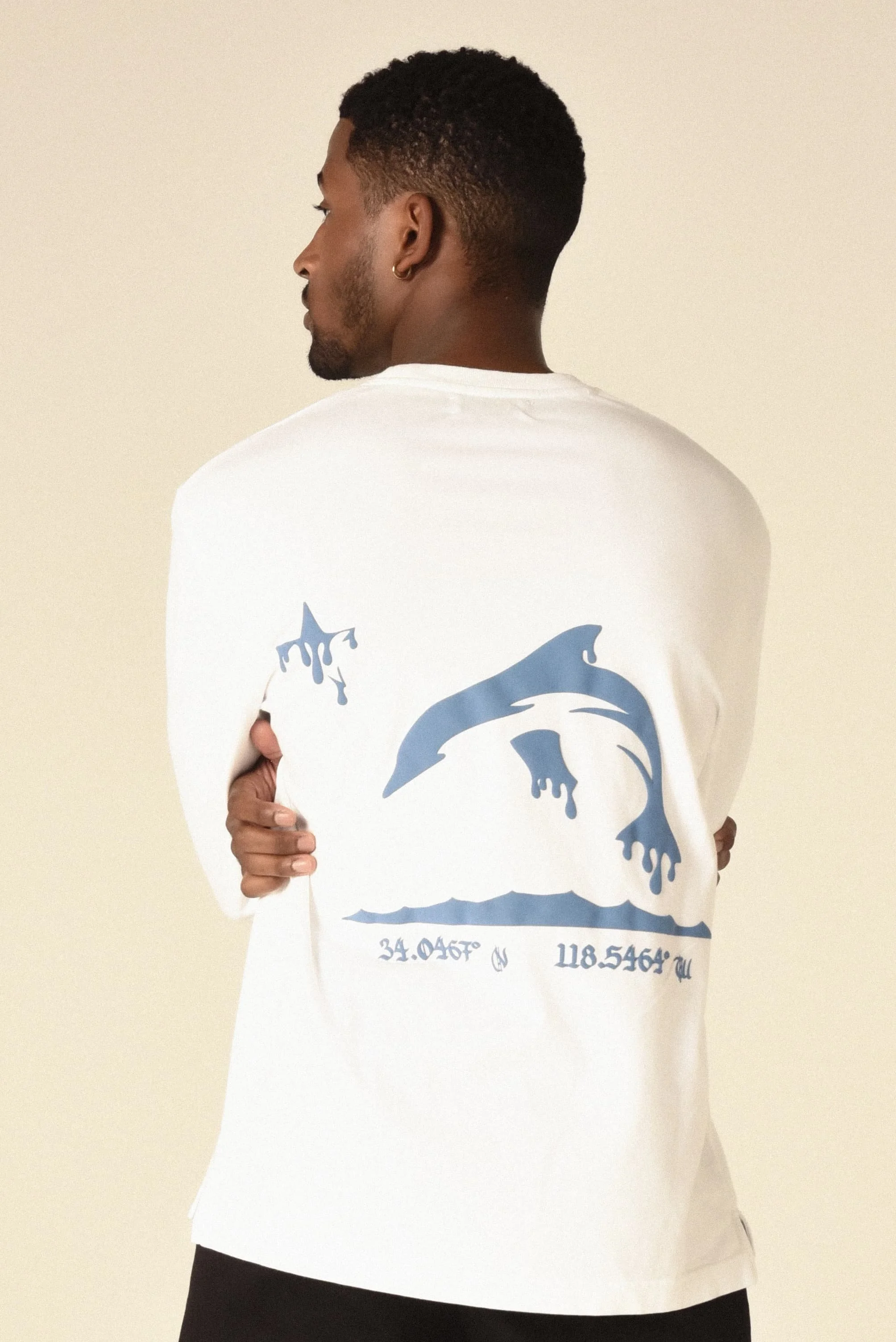 Model wearing a white T-shirt featuring a blue distressed dolphin graphic and geographic coordinates - designed in collaboration with Keagan Roberson.