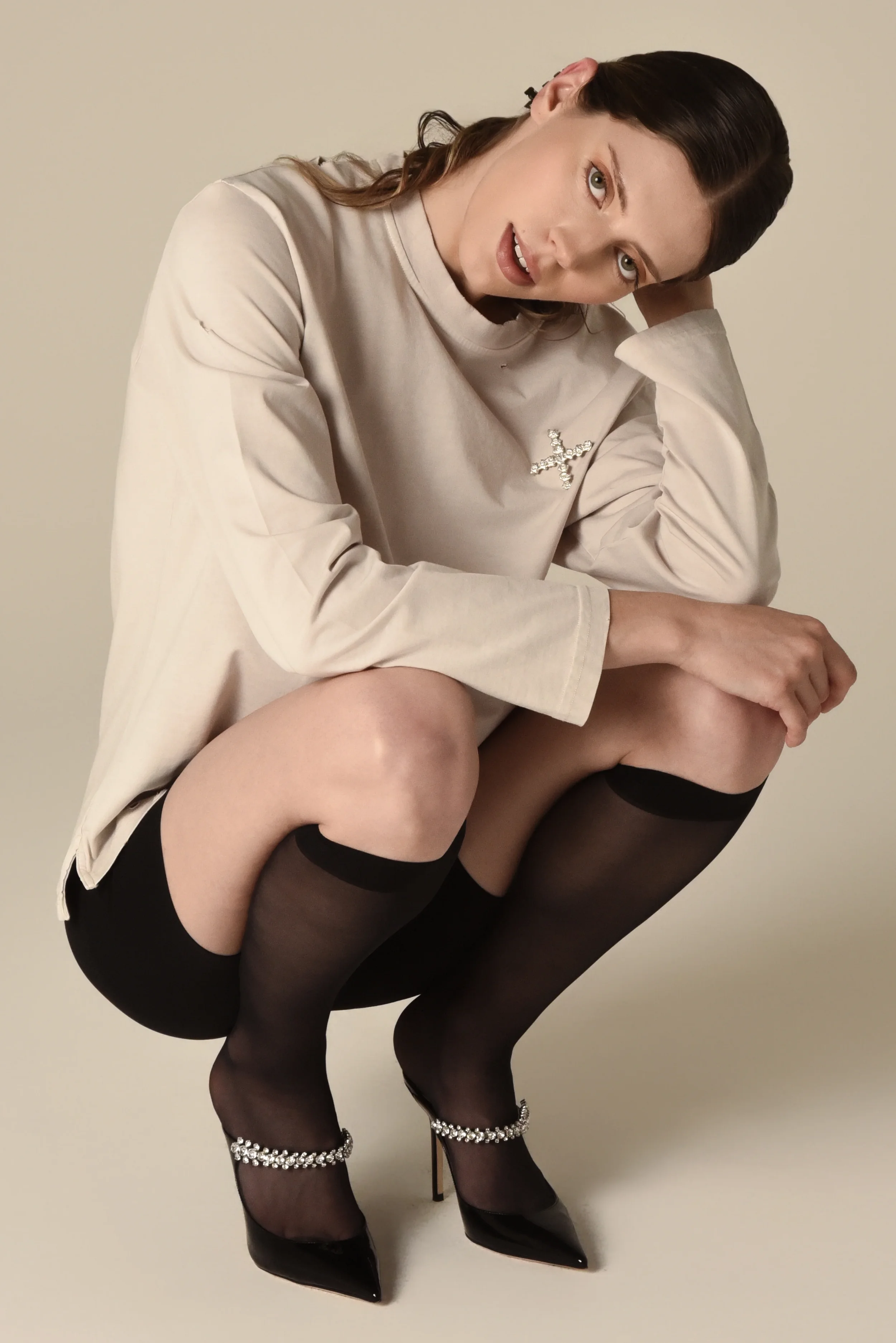 Model wearing a beige long-sleeve shirt with a rhinestone cross pin, styled with black thigh-high stockings and rhinestone-detailed heels - designed in collaboration with Keagan Roberson.