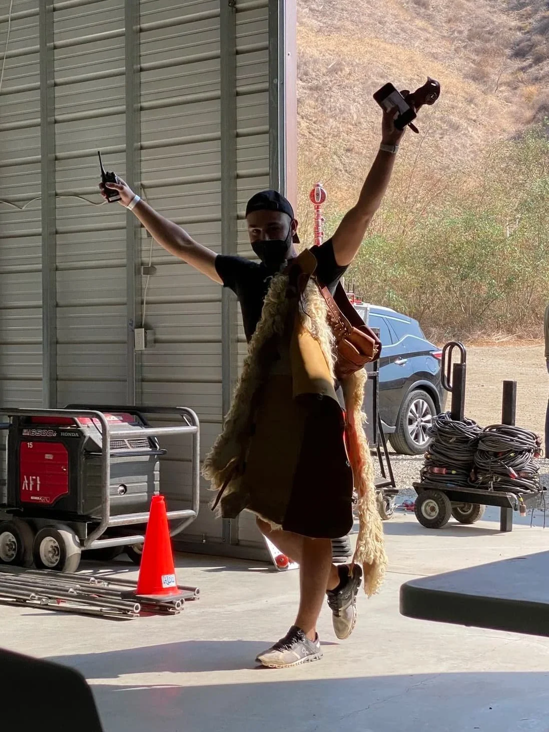 A person wearing a black mask, black T-shirt, and sneakers standing in a warehouse, raising both arms with a control in one hand and a camera in the other, in front of an open garage door with outdoor scenery visible.