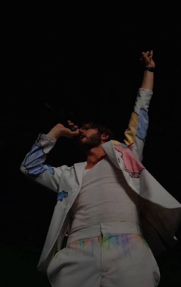 A person singing into a microphone with one arm raised, wearing a colorful, patched jacket and light-colored pants, against a black background.